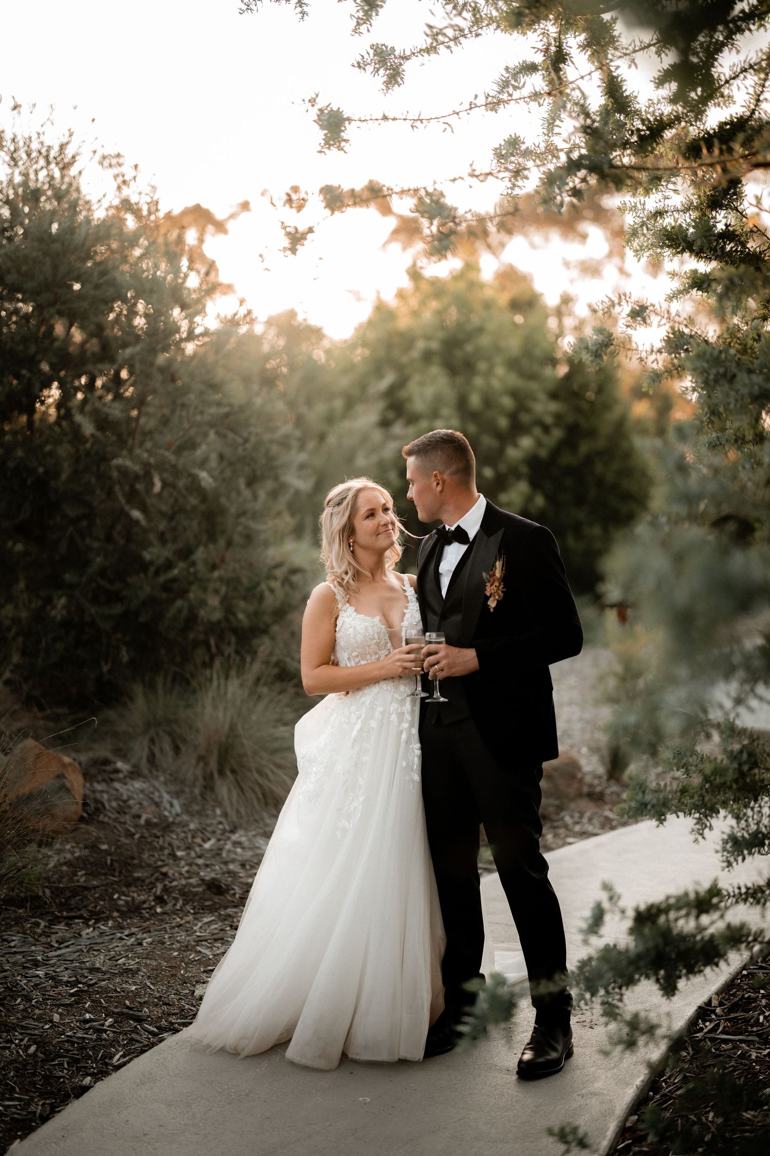 A newlywed couple poses on a pathway at sunset surrounded by trees and golden light.