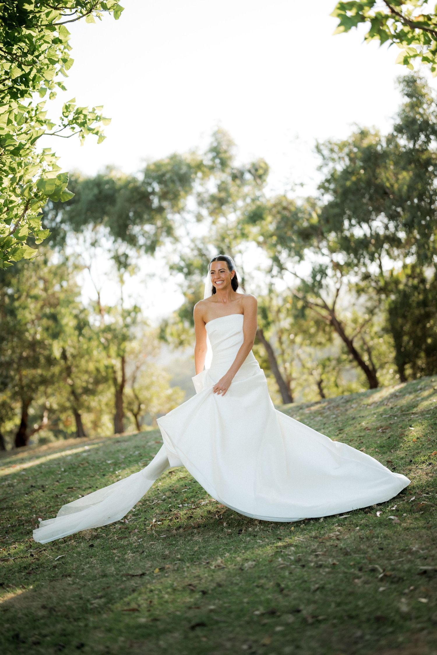 A flowing white wedding dress spreads across a grassy hillside in natural sunlight.
