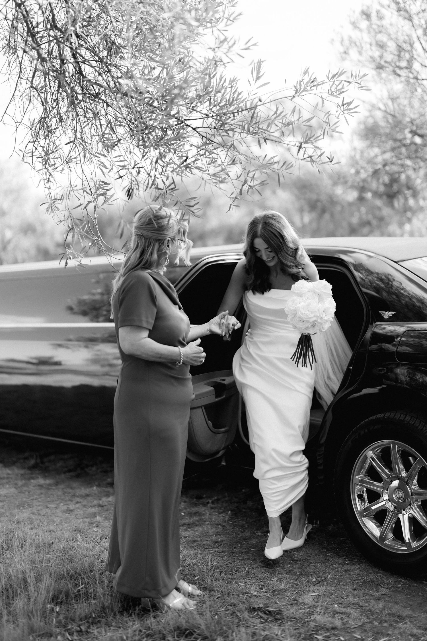 Black and white photo of bride getting assistance entering a luxury car.