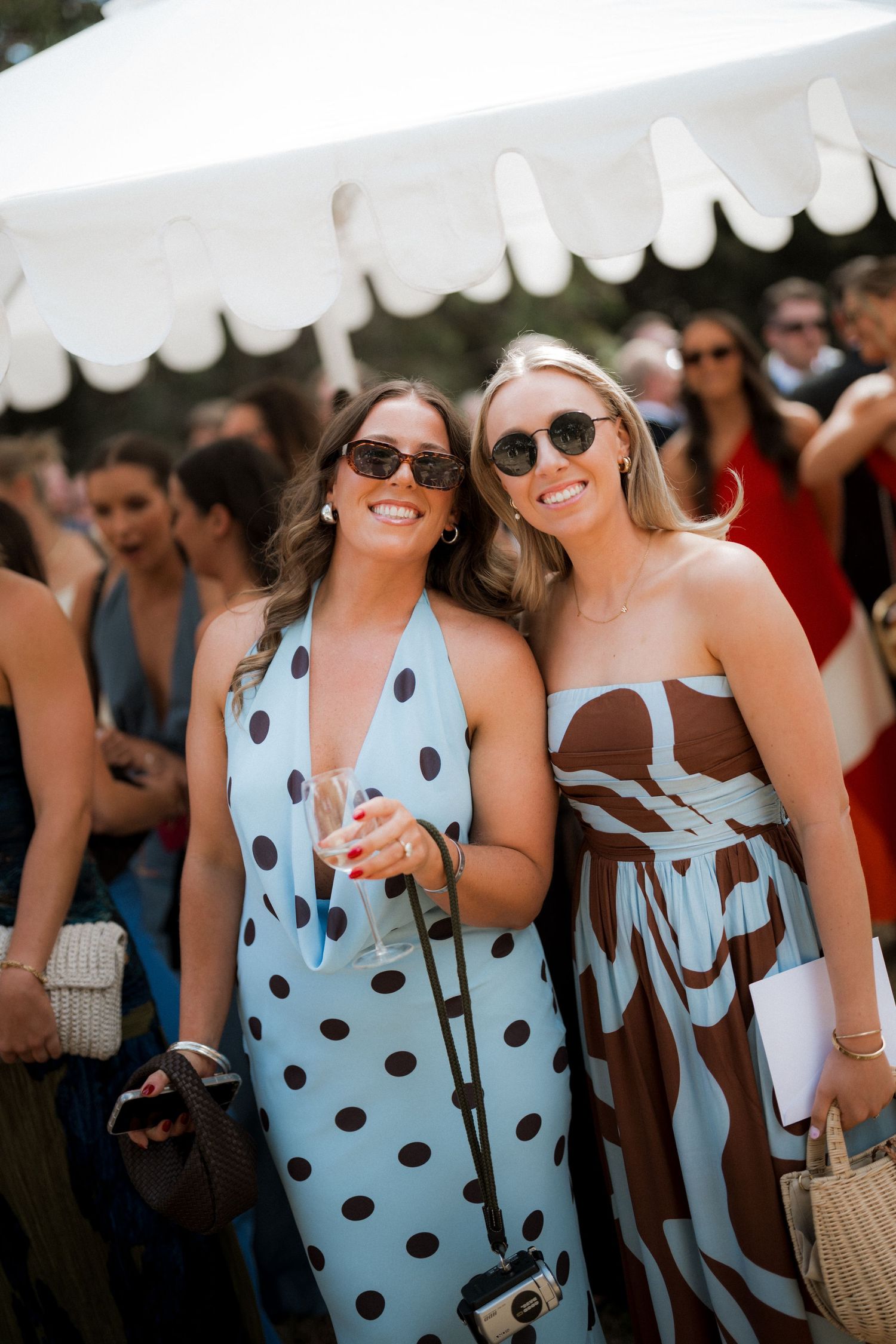 Two smiling women in stylish summer dresses posing together at an outdoor wedding reception under a white tent.
