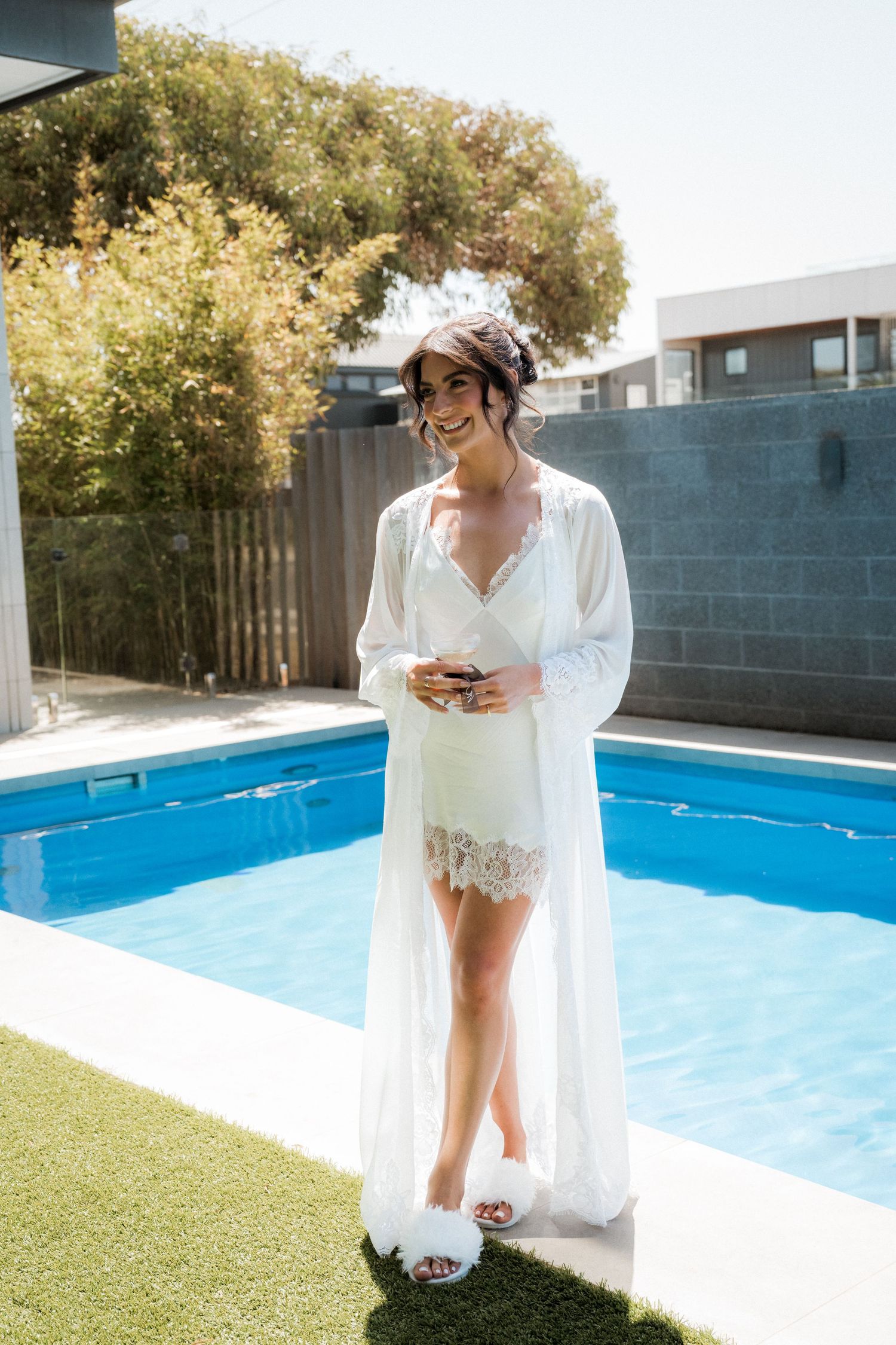 Bride in white robe poses by blue swimming pool on sunny day.