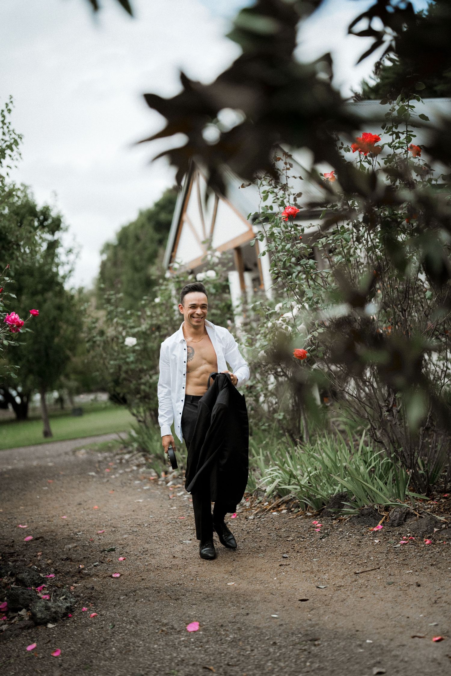 Smiling groom walks garden path in open white shirt carrying black jacket amid rose bushes and green trees.