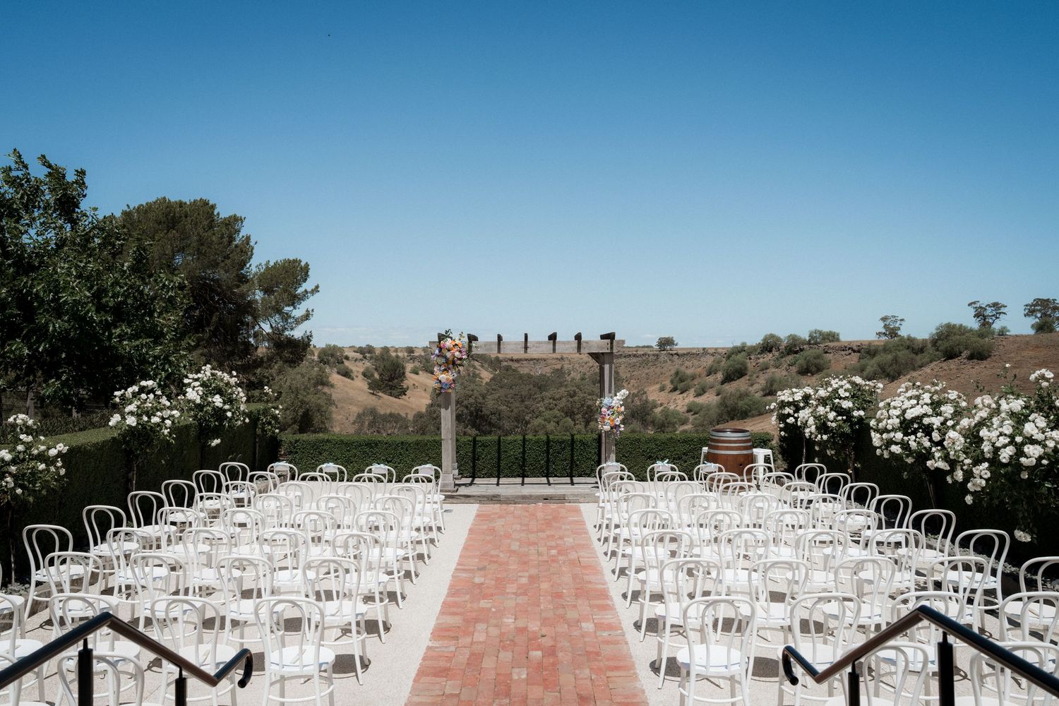 Elegant outdoor wedding ceremony setup with white chairs, brick aisle, floral arrangements, and clear blue sky backdrop.