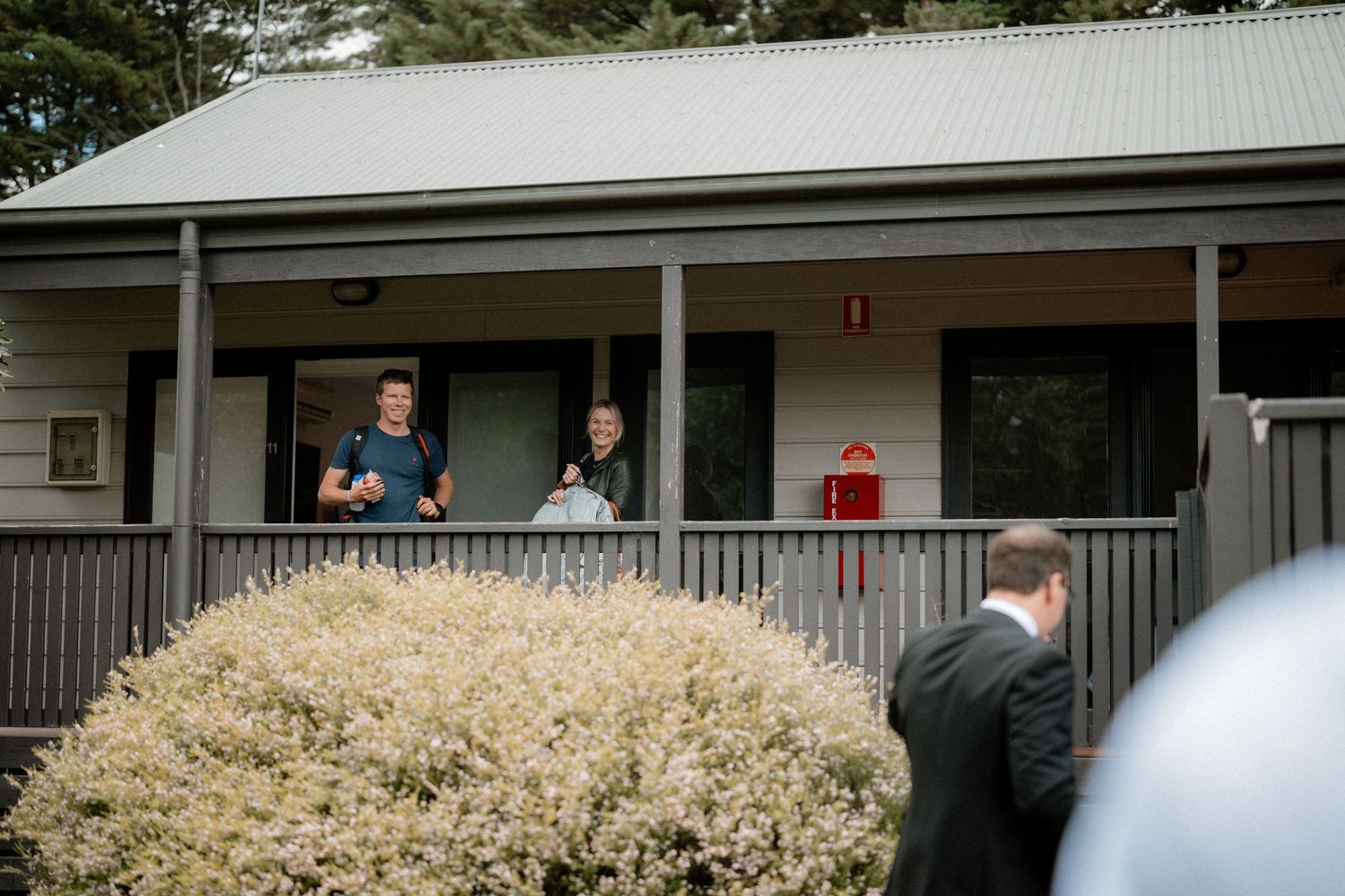 People stand on a covered porch of a rustic building with metal roof and bushes in the foreground.