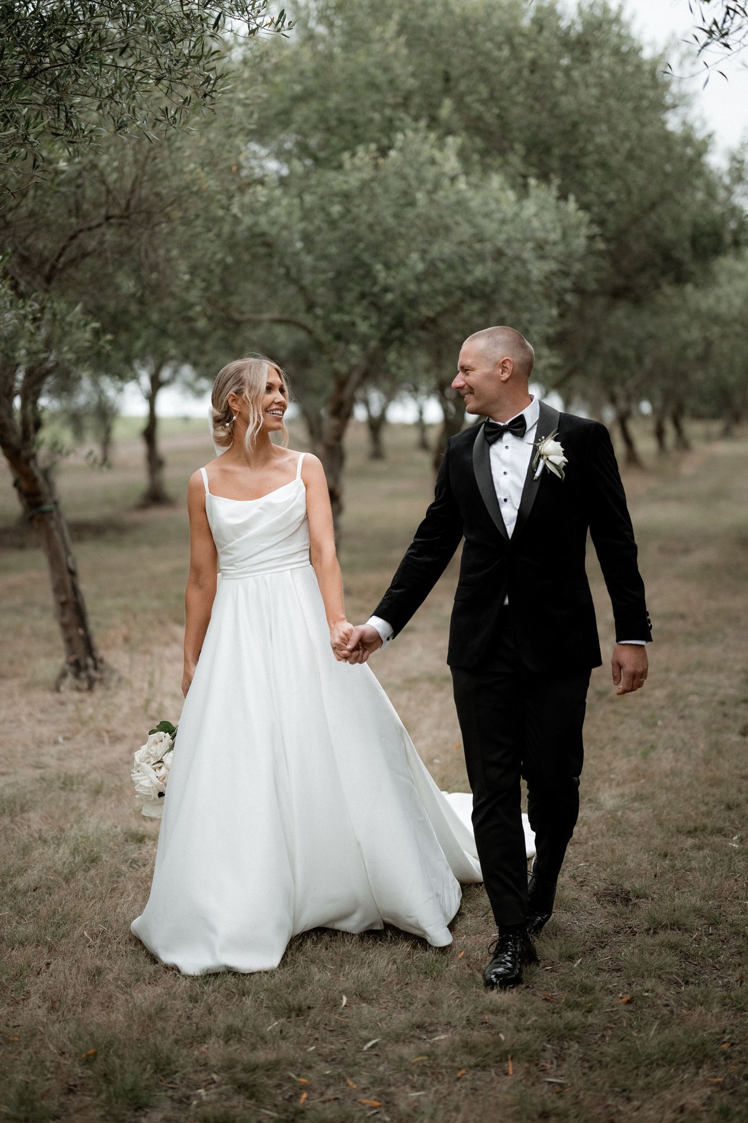 Newlyweds share joyful moment walking through olive grove in formal wedding attire.