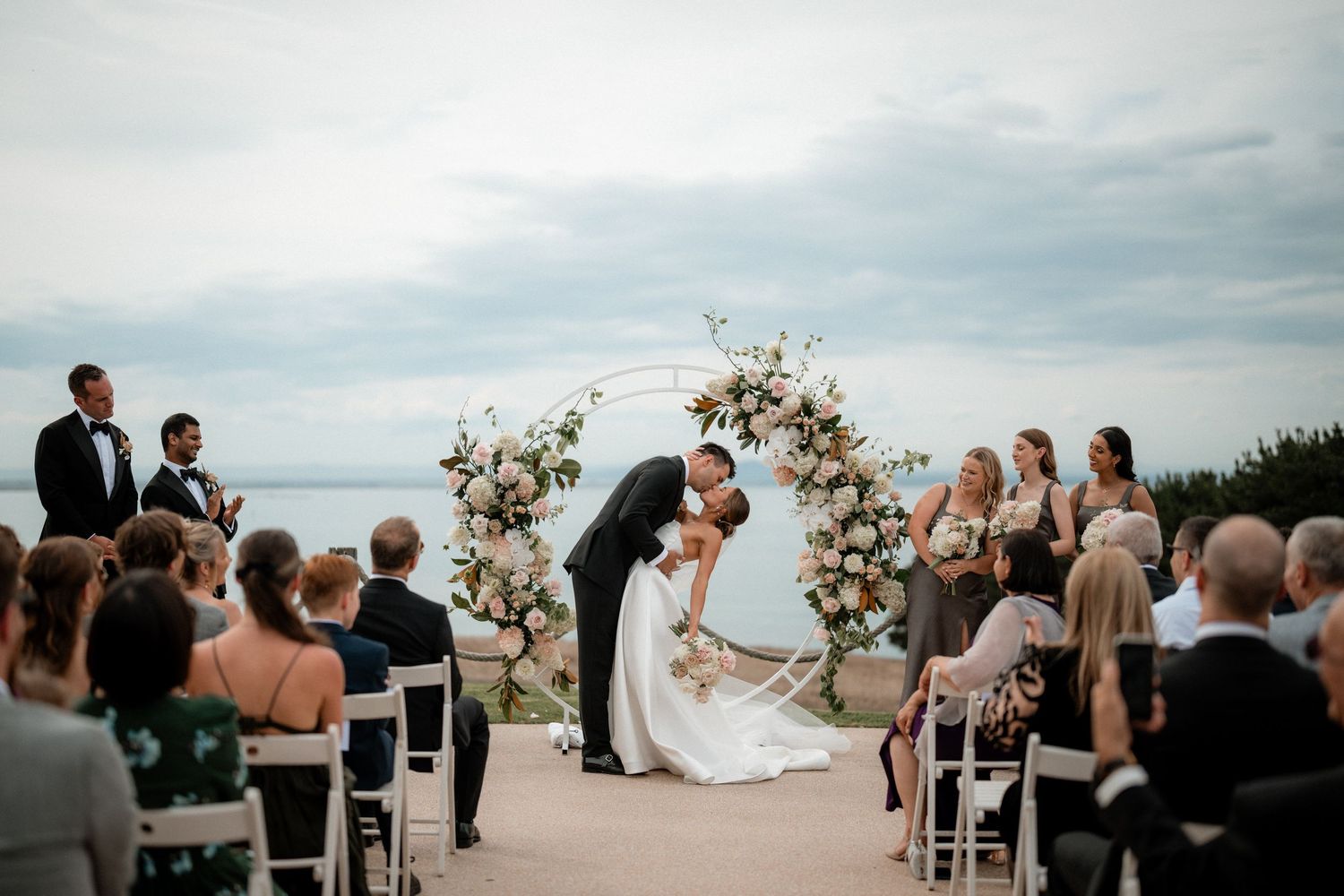 A wedding ceremony takes place outdoors with guests seated in white chairs facing an arch decorated with floral arrangements.