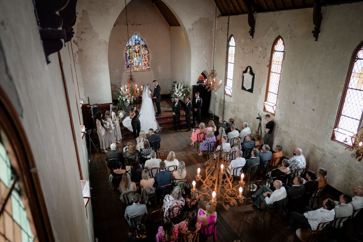 An aerial view of a wedding ceremony taking place inside a historic chapel with stained glass windows and gathered guests.