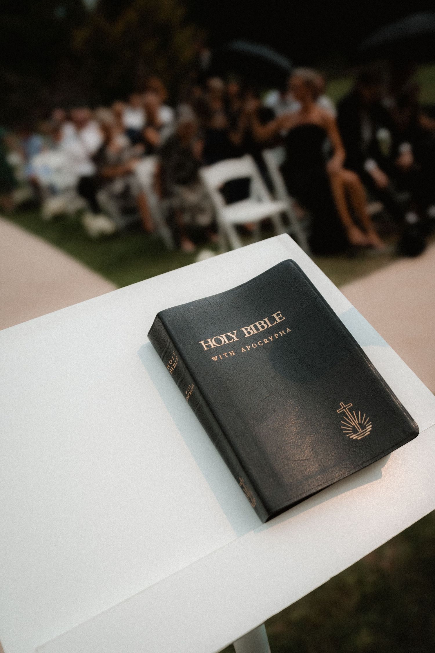 A black Holy Bible rests on a white surface during an outdoor gathering with blurred guests in the background.