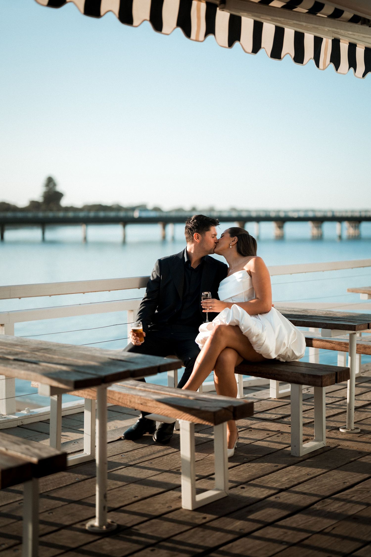 A romantic couple in wedding attire share an intimate moment on a waterfront pier under a striped awning.