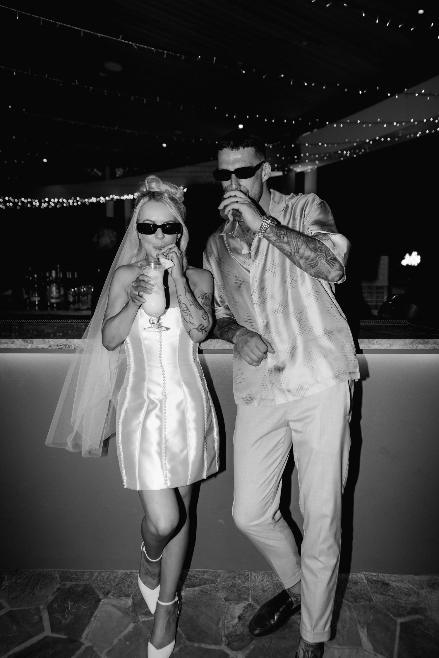 Couple in white attire sharing drinks under string lights at night.