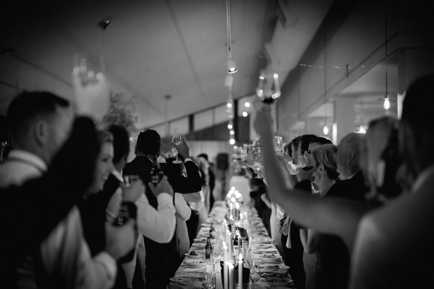 Black and white photo of wedding guests raising glasses in celebration along a banquet table.