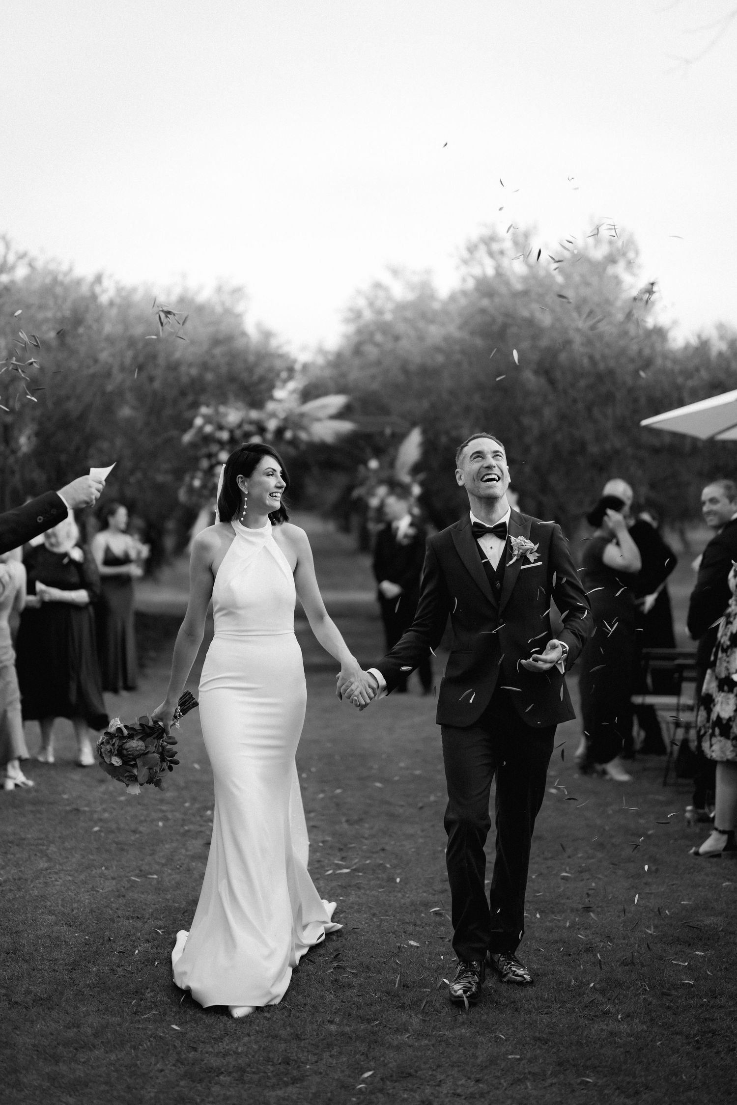 A black and white photo of a couple celebrating at their outdoor wedding while guests throw confetti.