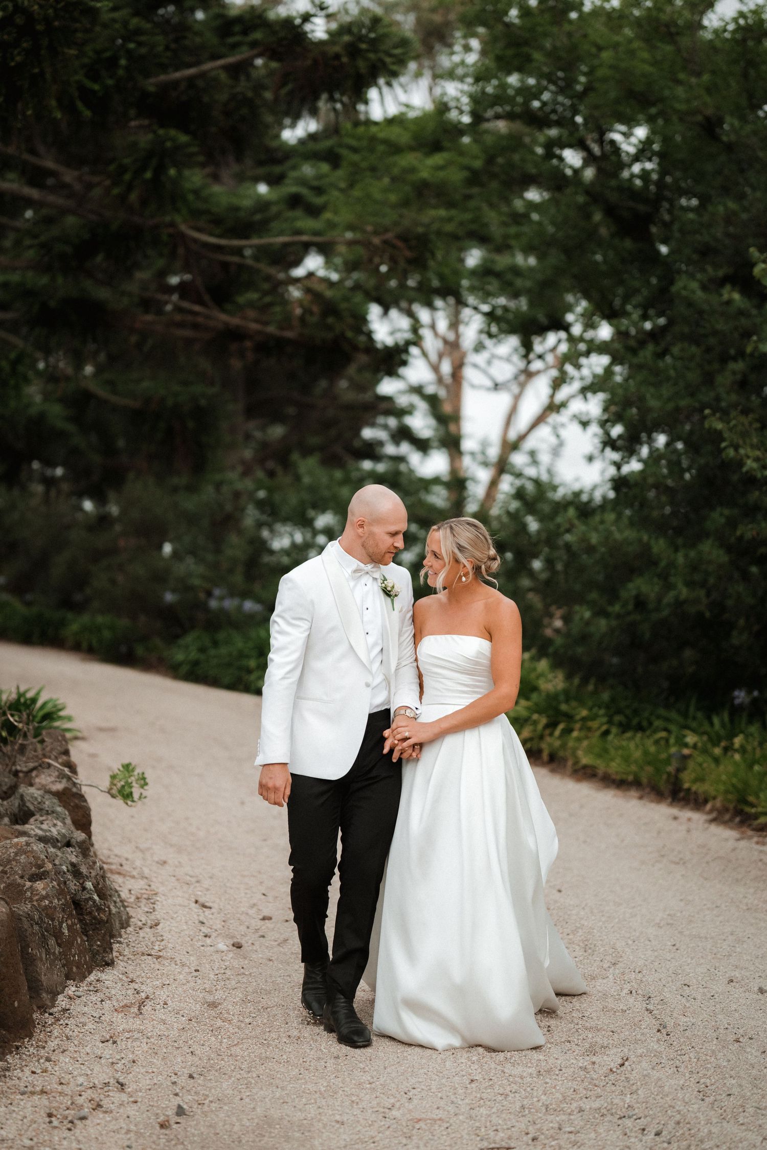 Elegant couple walks together on a garden path surrounded by towering pine trees.