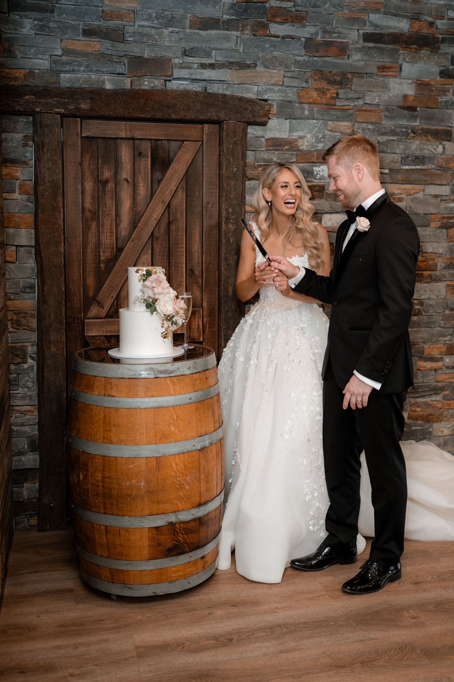 A wedding couple laughs together next to their cake displayed on a rustic wine barrel.