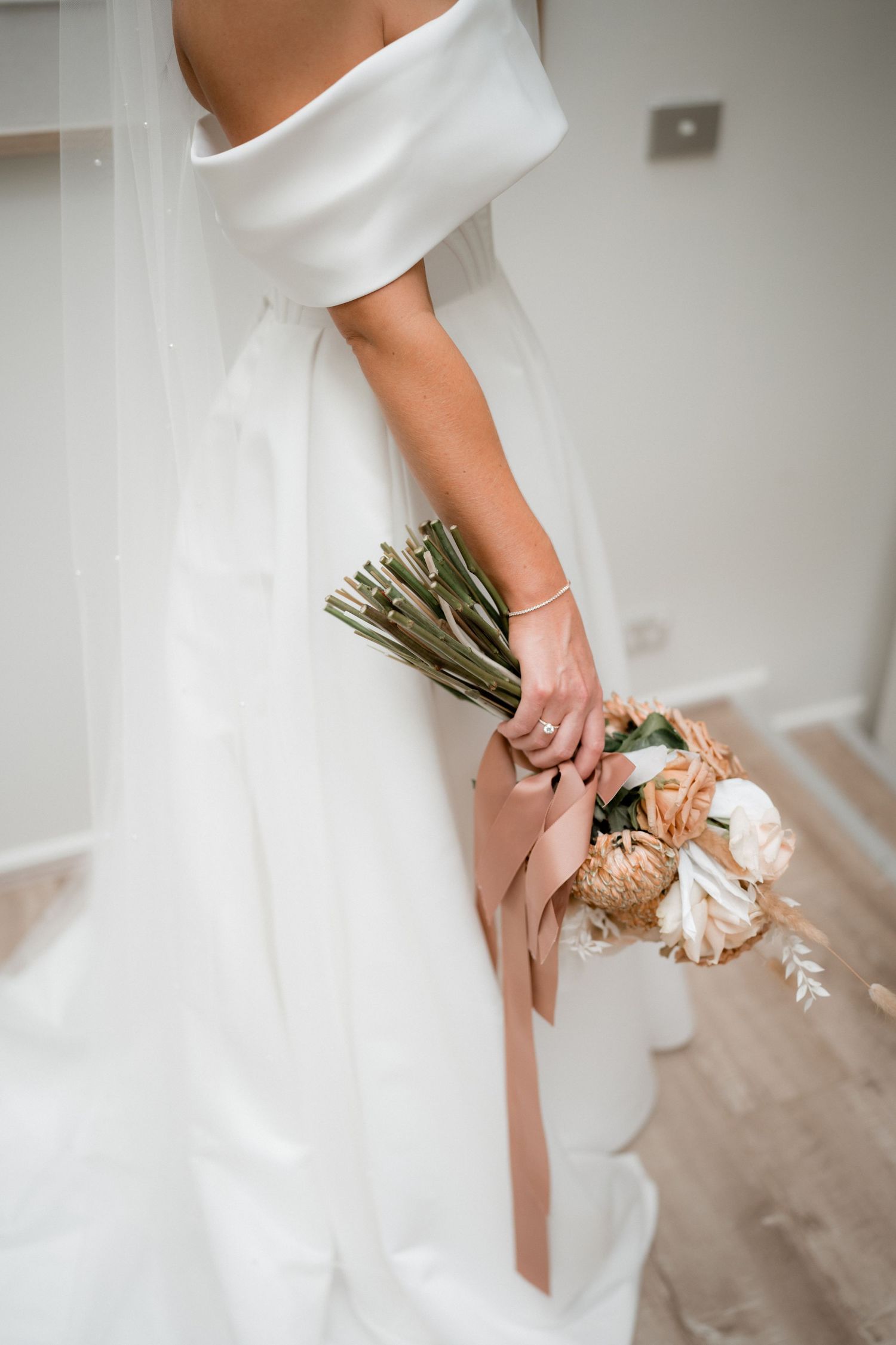 Close-up view of bride in white dress holding a delicate bouquet with a blush pink ribbon against a neutral wall.