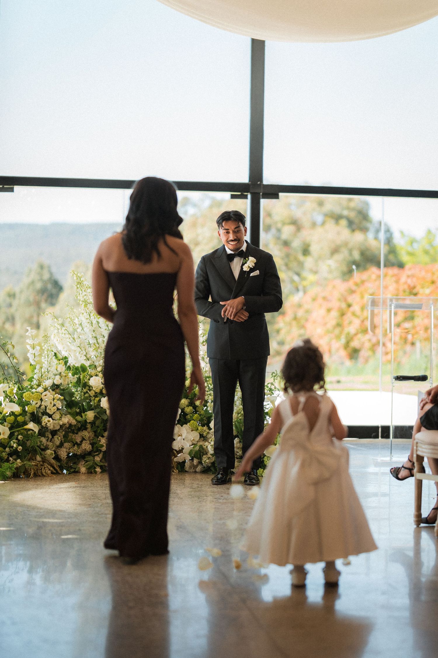 Groom in a black tuxedo smiles from the aisle as a bridesmaid and a flower girl in a white dress walk toward him at a wedding ceremony.