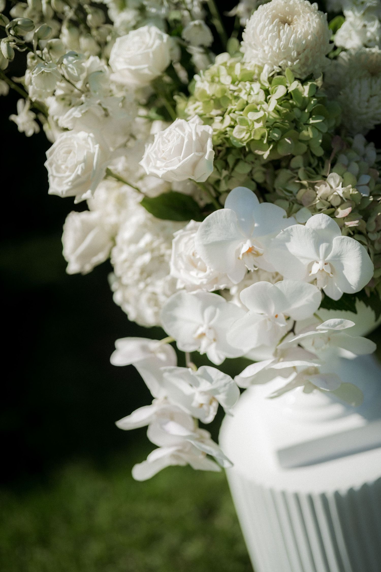 White hydrangea and rose floral arrangement in a classic white pedestal vase against a dark background.
