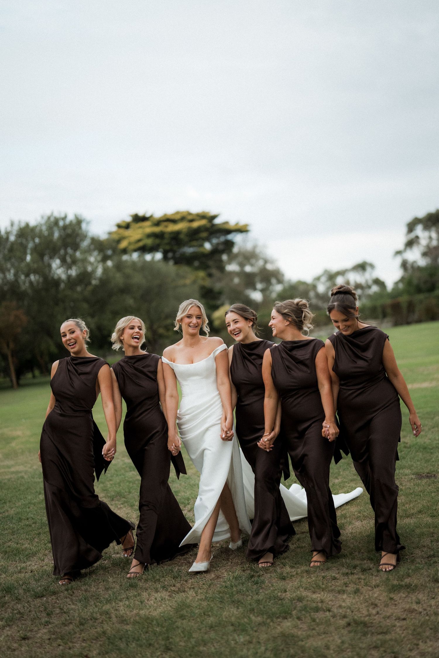 Wedding party performs synchronized leaning pose on grassy hill in formal attire.