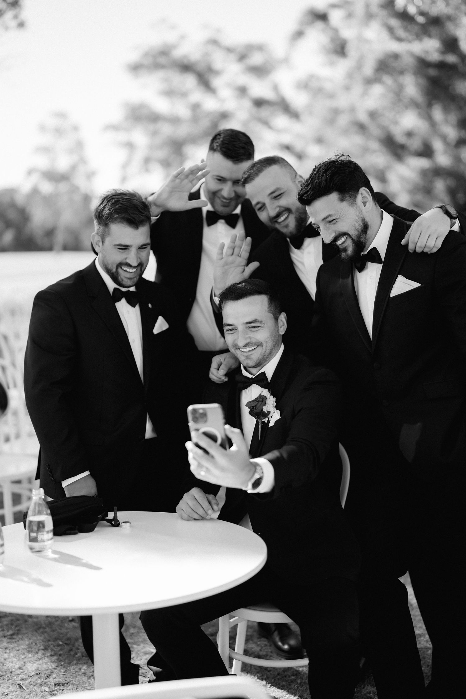Wedding party members in formal attire sharing laughs around a cocktail table outdoors.