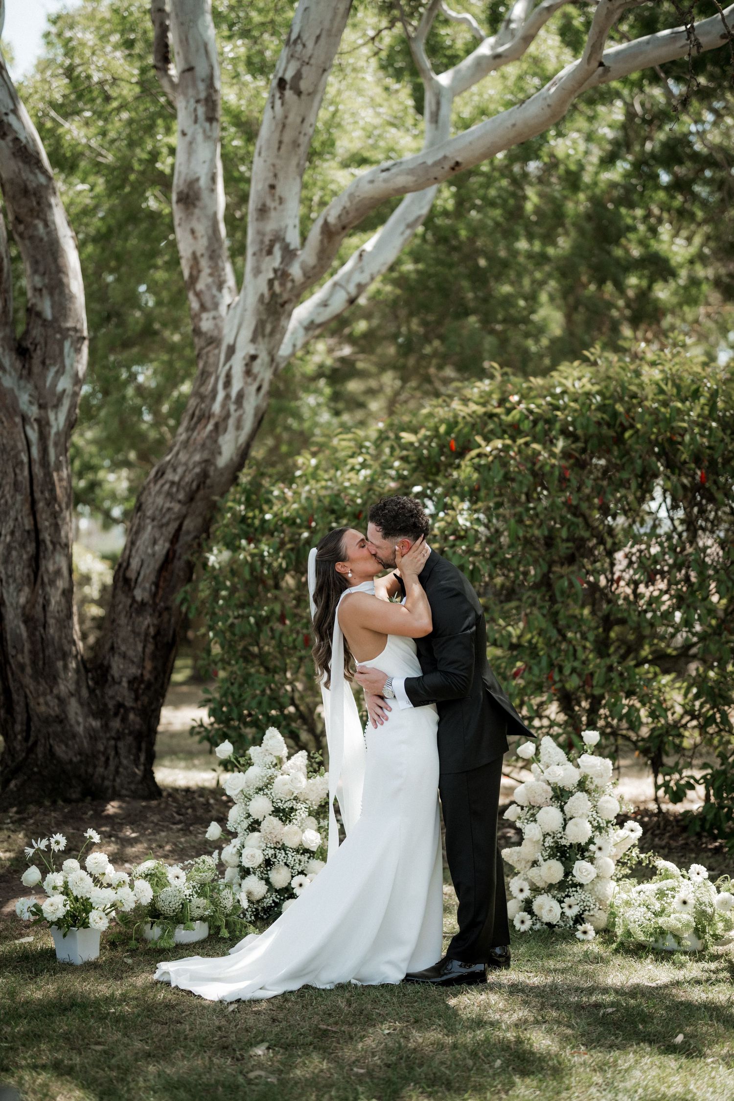 A romantic wedding couple embraces under tall eucalyptus trees with white floral arrangements on the ground.