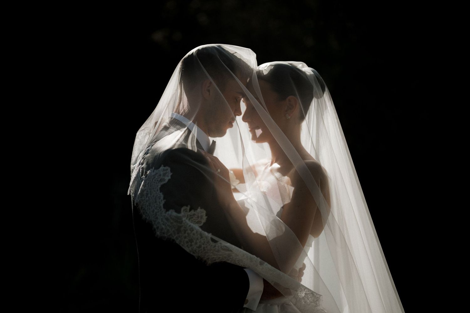 A romantic silhouette of a couple embracing under a flowing white veil in dramatic lighting.