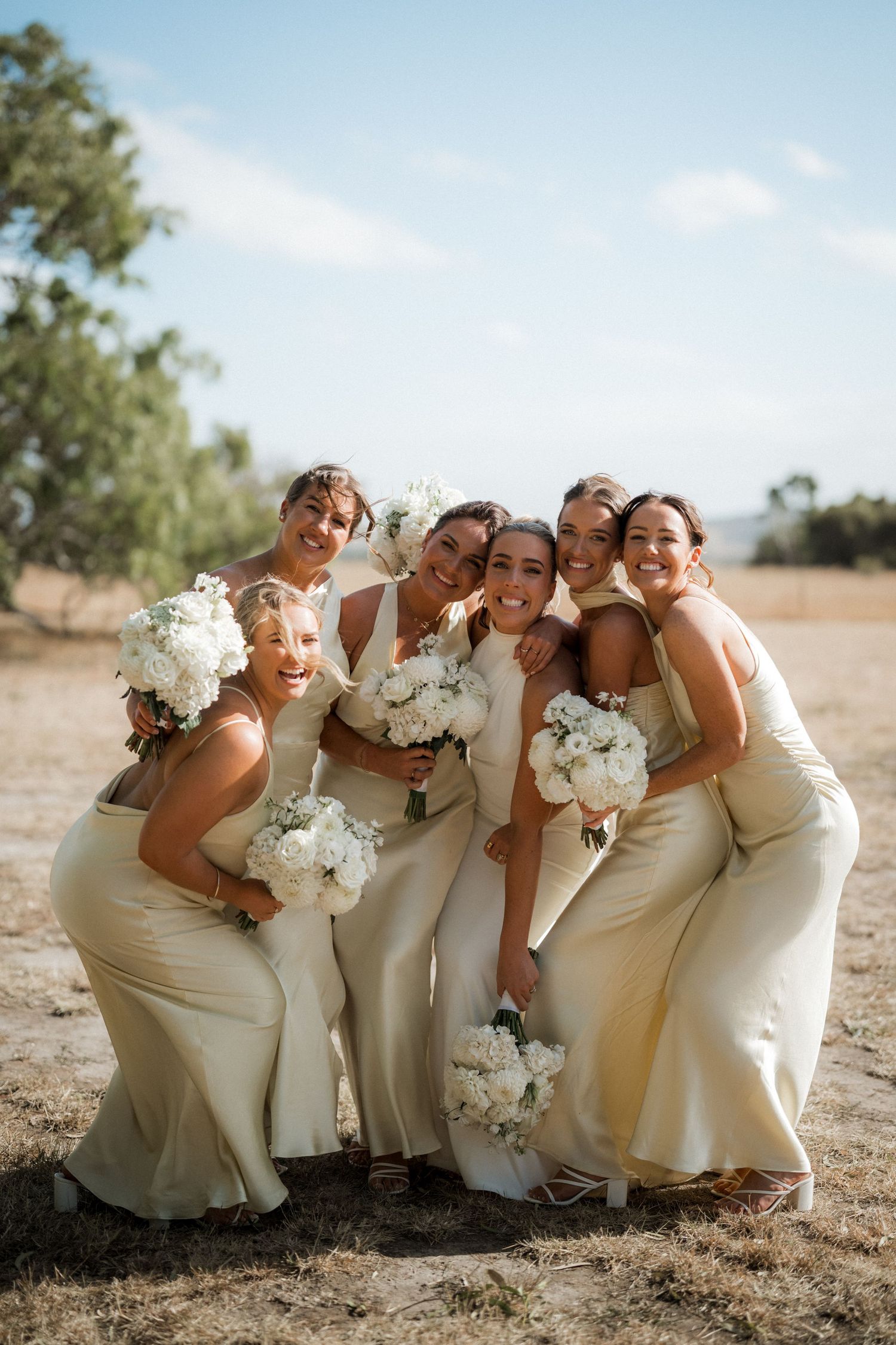 Bridesmaids in cream-colored dresses posing playfully with bouquets in an outdoor setting.
