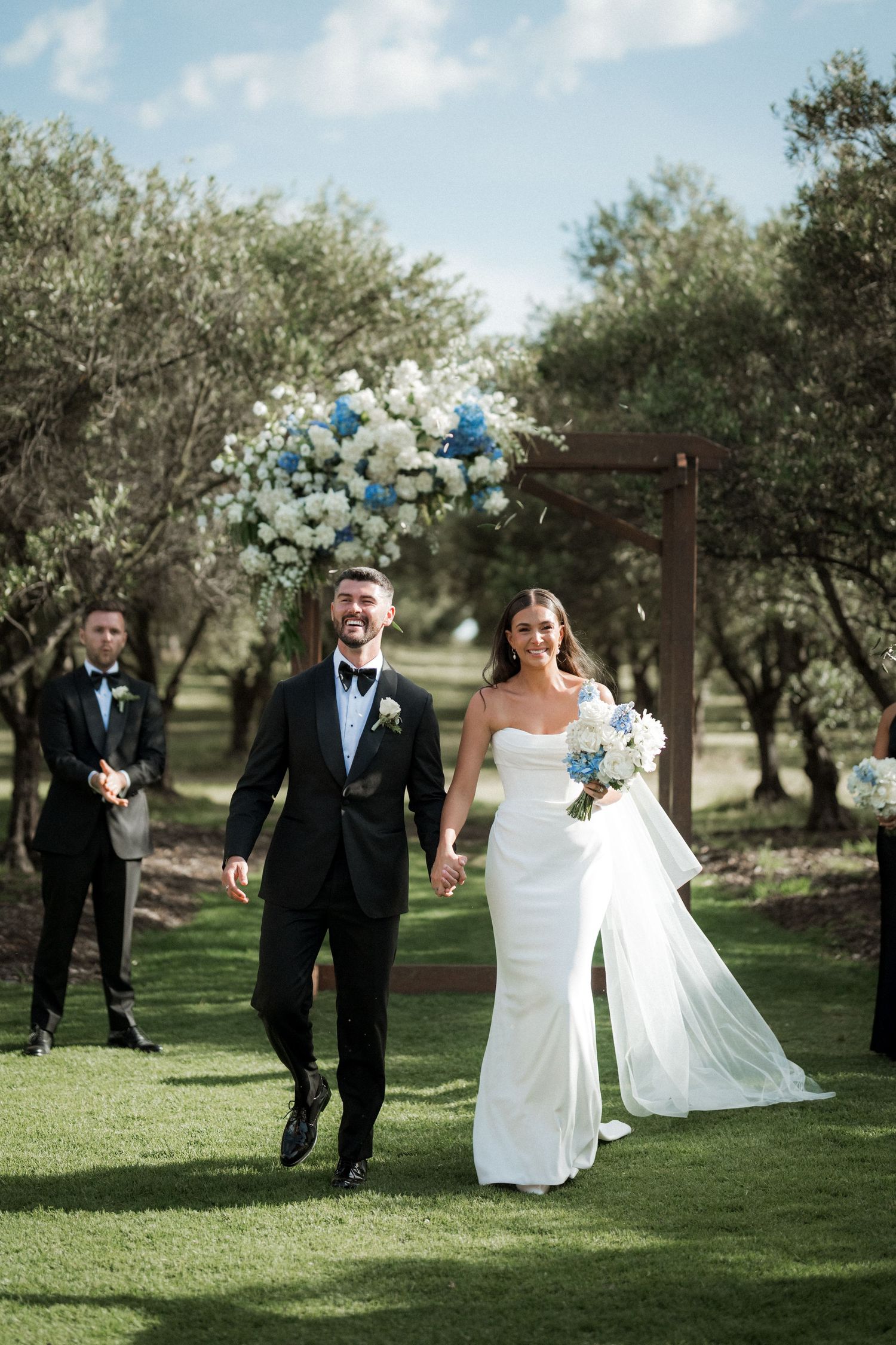 Wedding party walks and poses together on green lawn with olive trees in background.