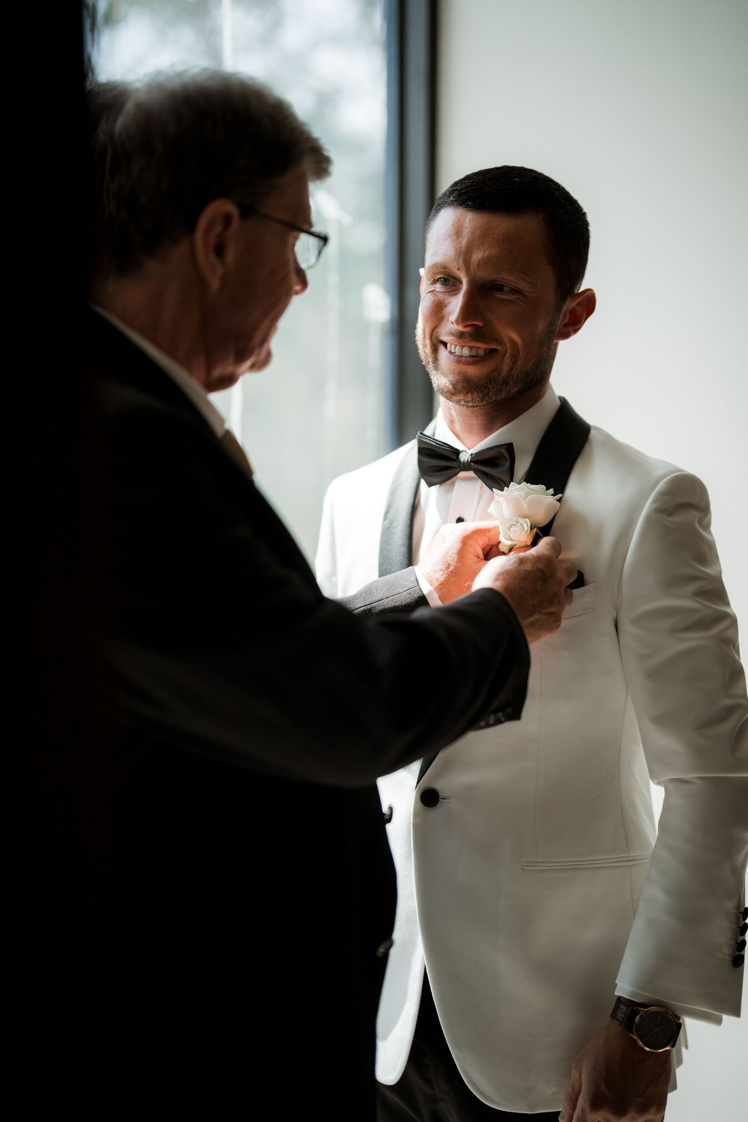 A formal scene showing someone helping adjust a bowtie on a person wearing a white tuxedo jacket.