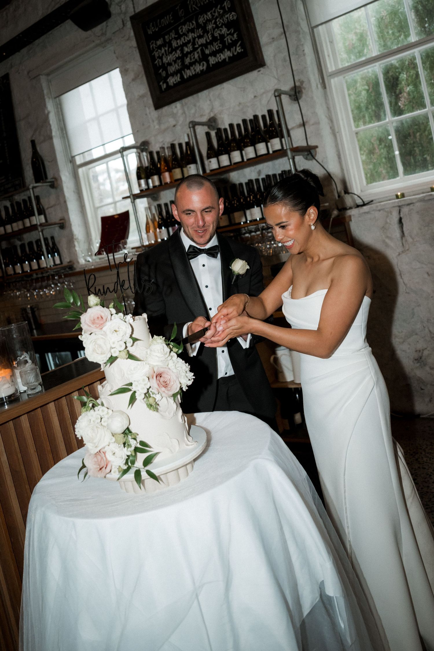 Wedding couple cutting their white floral wedding cake together in a rustic indoor venue.