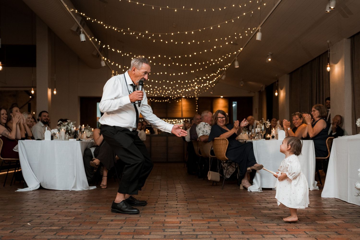 Wedding reception venue with string lights and guests dancing on wooden floor.