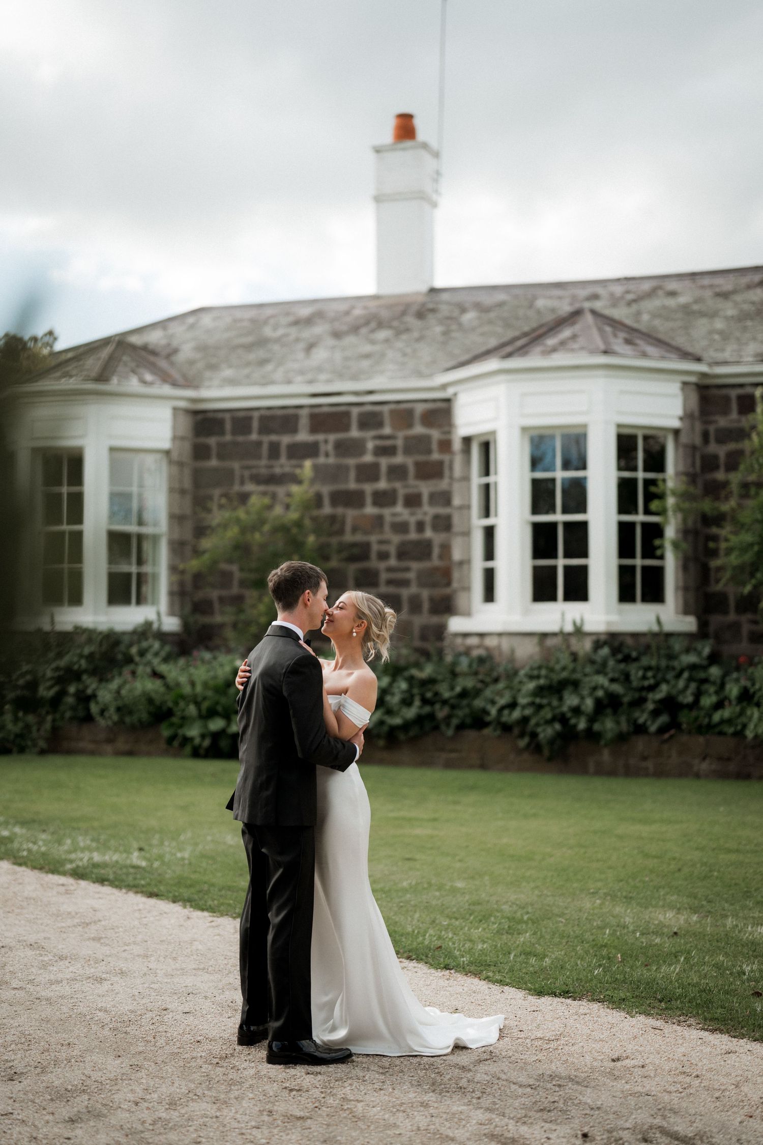 Couple poses in front of a stone house with white trim and bay windows.