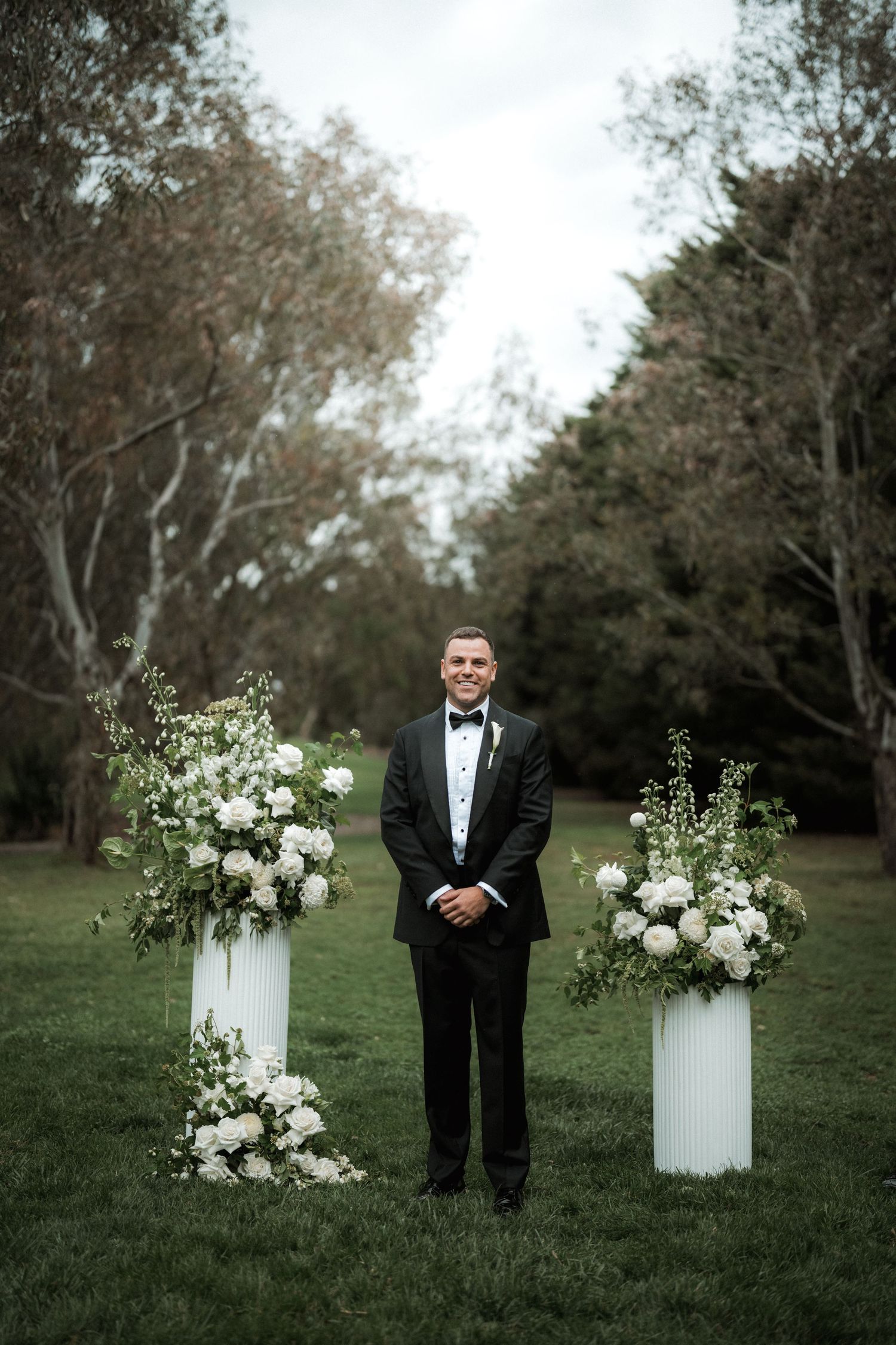 Elegant white floral arrangements on pedestals line a garden path with formal attire.