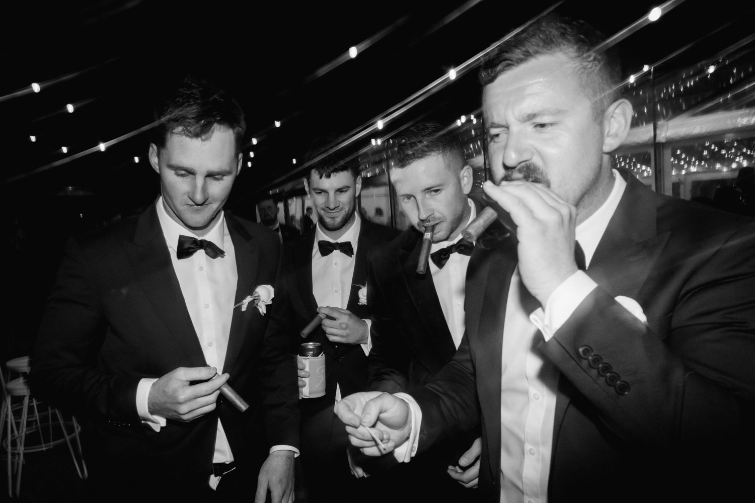 Black and white photo of groomsmen in tuxedos smoking cigars and drinking at a wedding reception.
