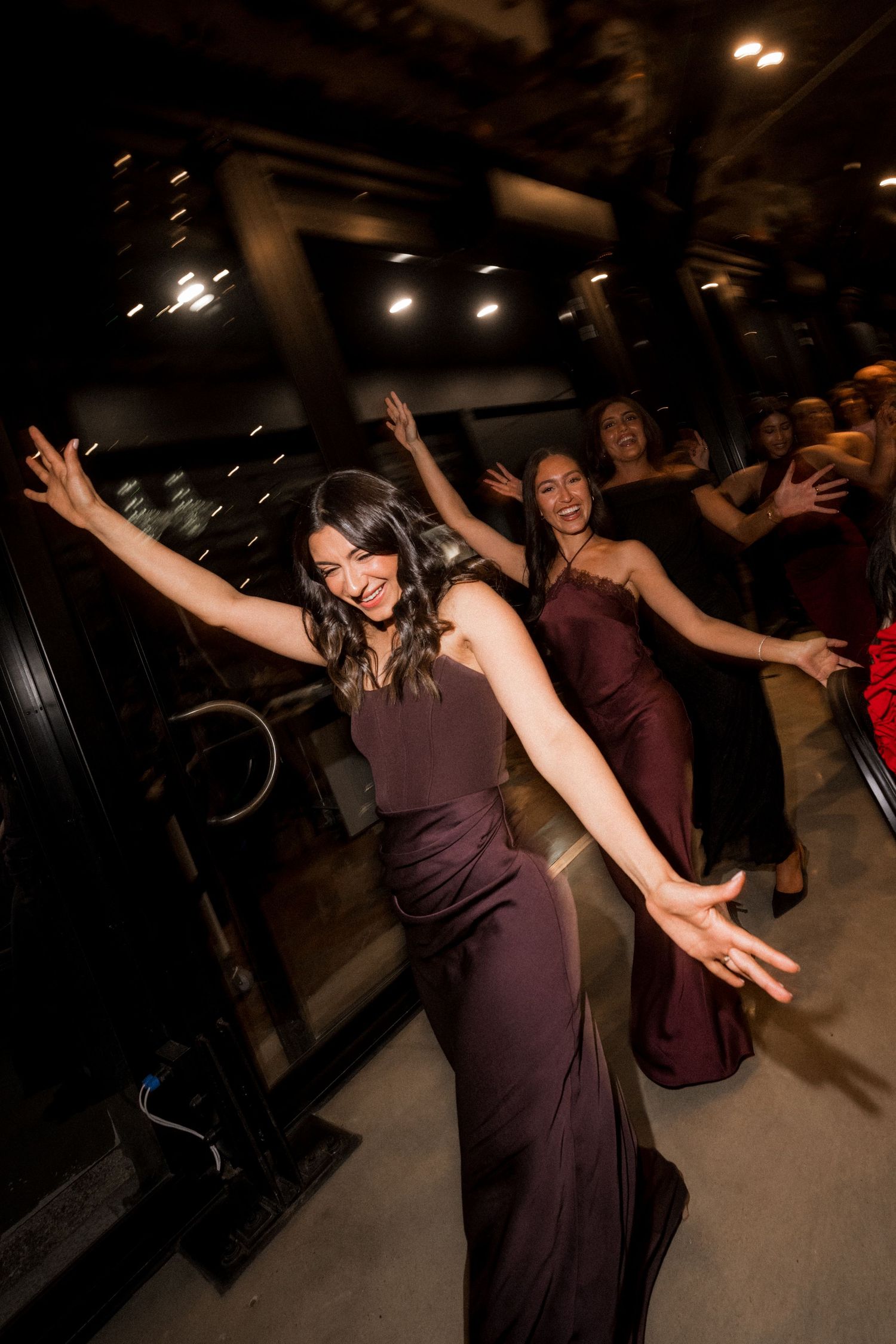 Bridesmaids in burgundy satin dresses dance joyfully with arms outstretched at an evening wedding reception.