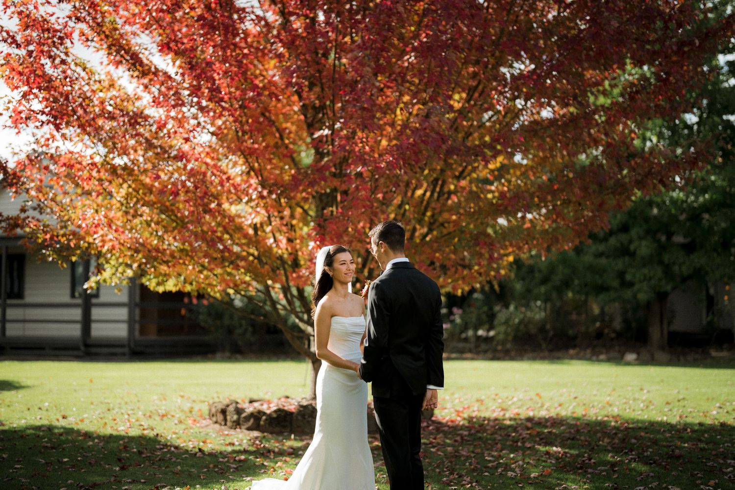 Bride and groom share a romantic moment under a vibrant red autumn tree on lush green grass.