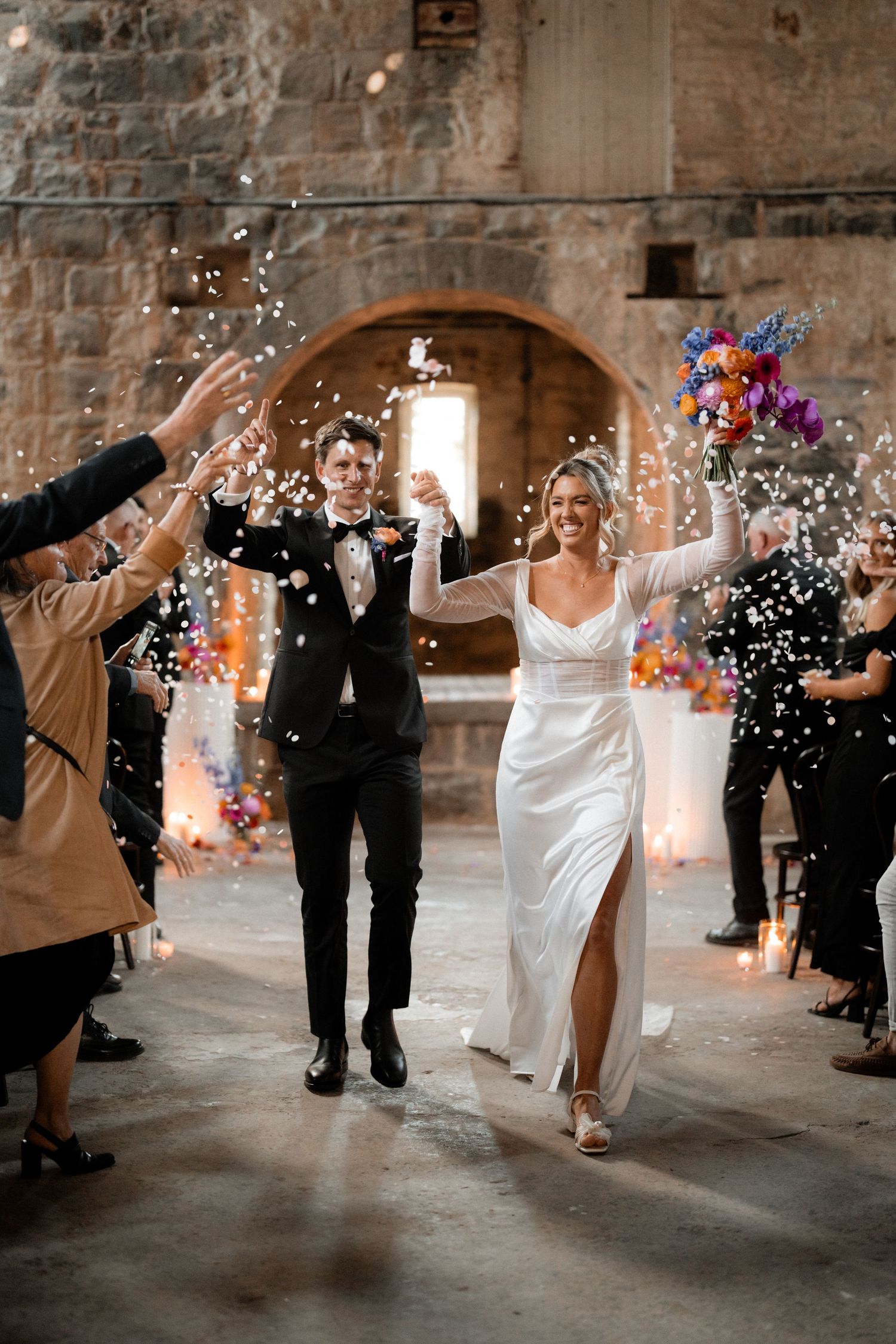 Newlyweds walk hand in hand through a stone archway while guests throw confetti during a wedding celebration.