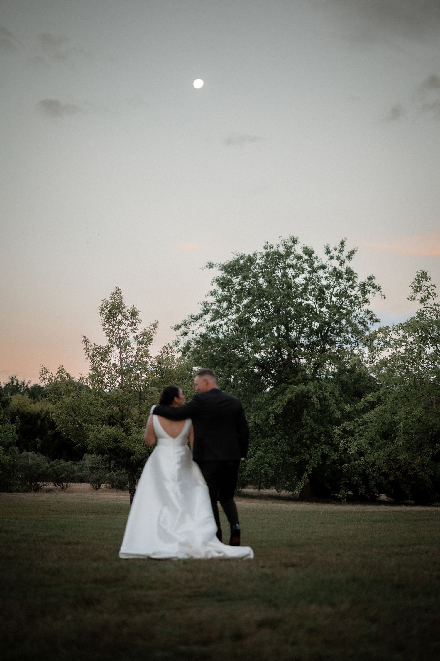 A couple stands together on a grassy field at dusk with a full moon glowing in the twilight sky.