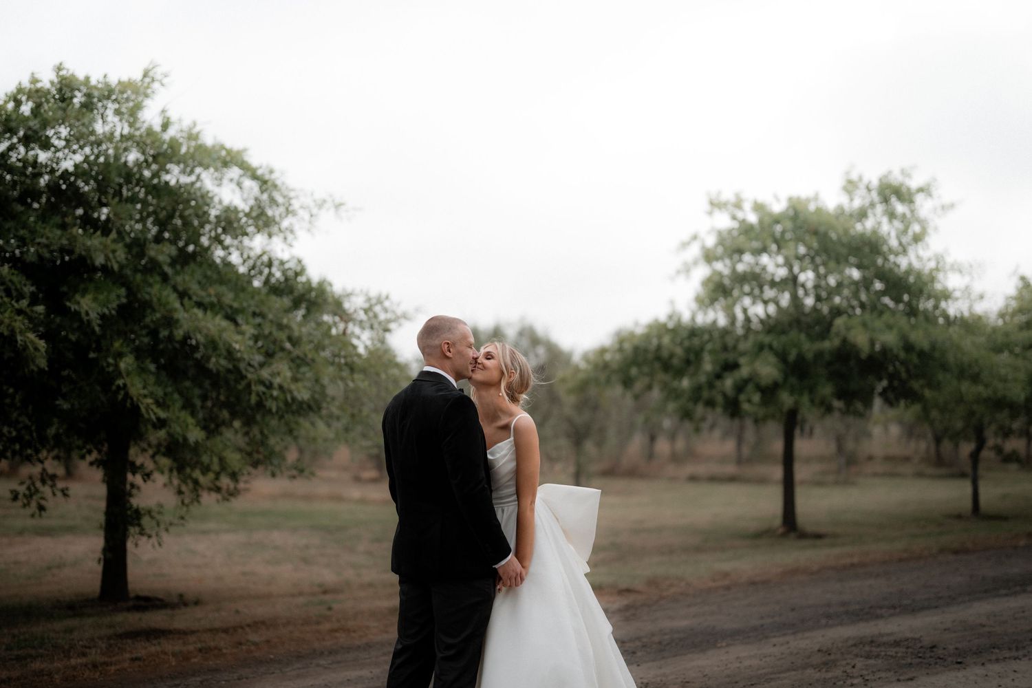A romantic moment captured between a couple in formal attire standing among orchard trees at dusk.