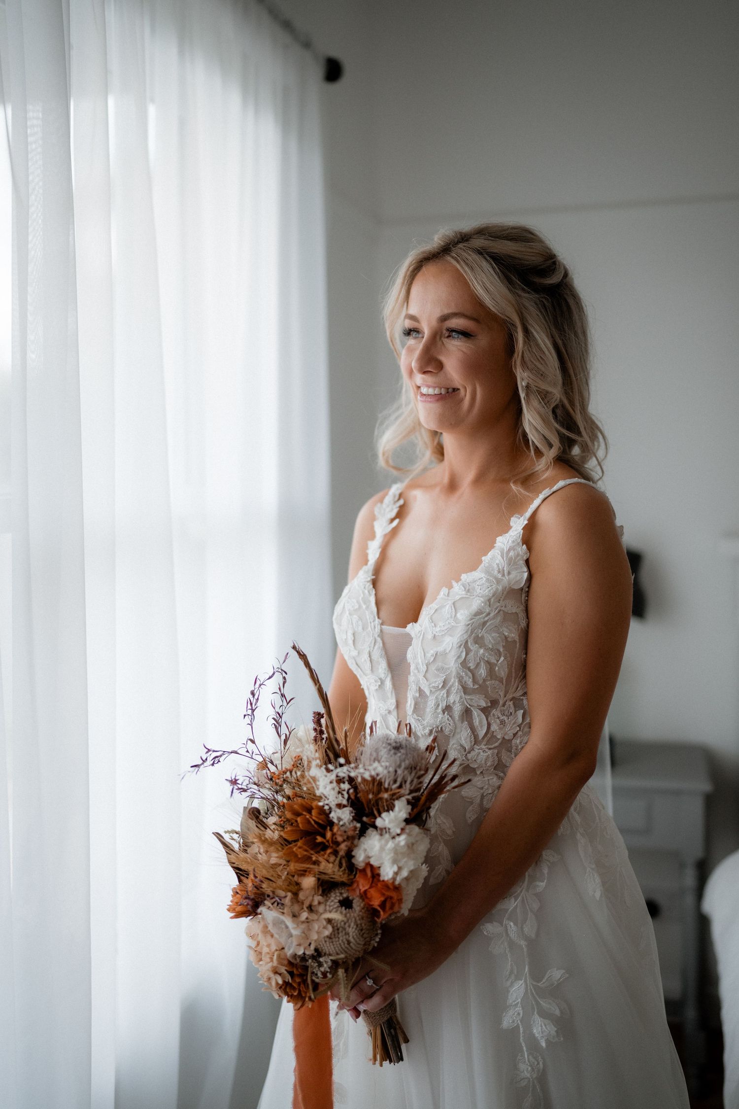 A bride in a white lace wedding gown stands by a window holding a bouquet of dried autumn flowers.