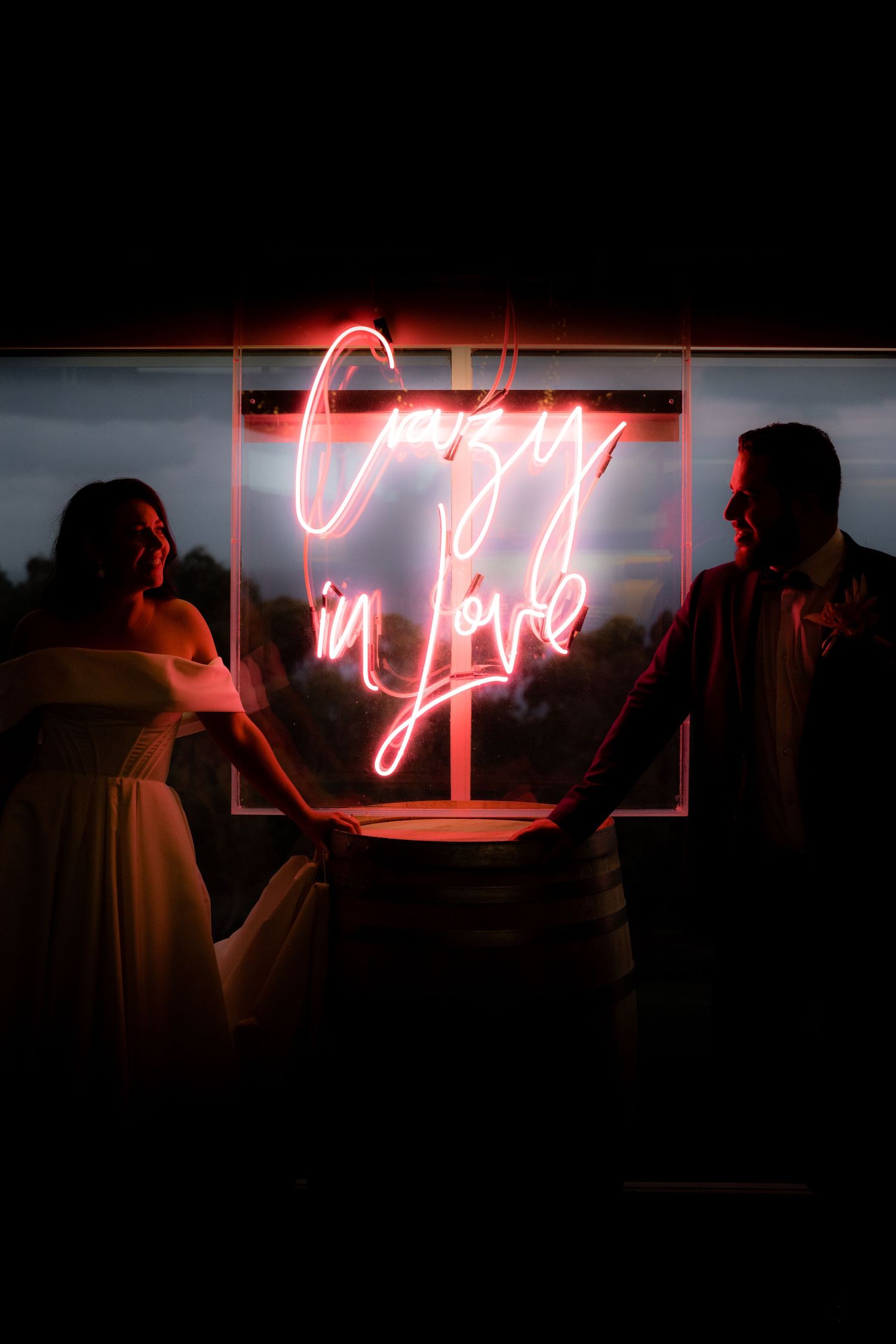 Silhouettes of two people stand in front of a glowing red neon 'crazy in love' sign in the darkness.