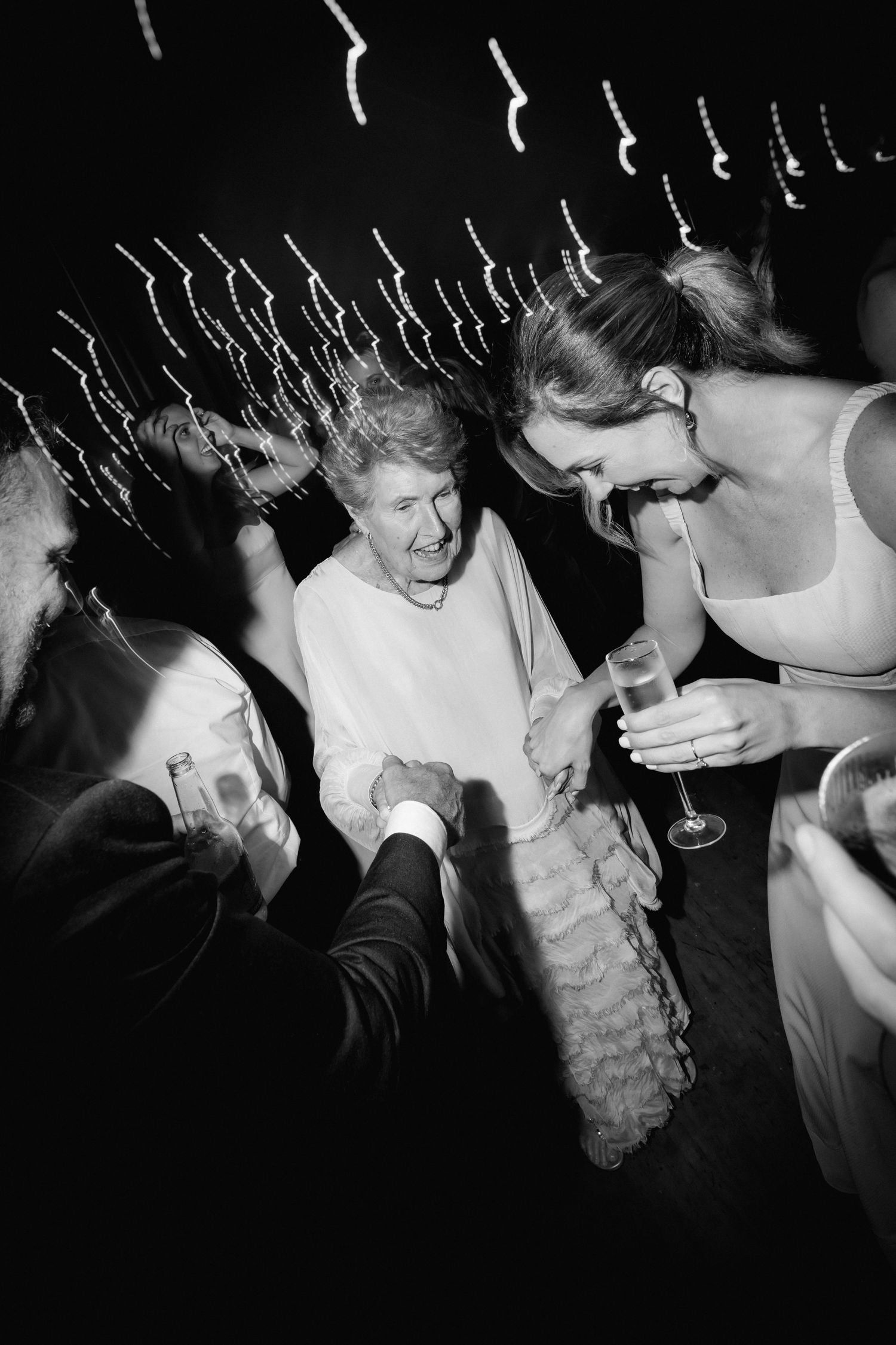 Black and white party photo showing wedding guests dancing and celebrating with drinks under string lights.
