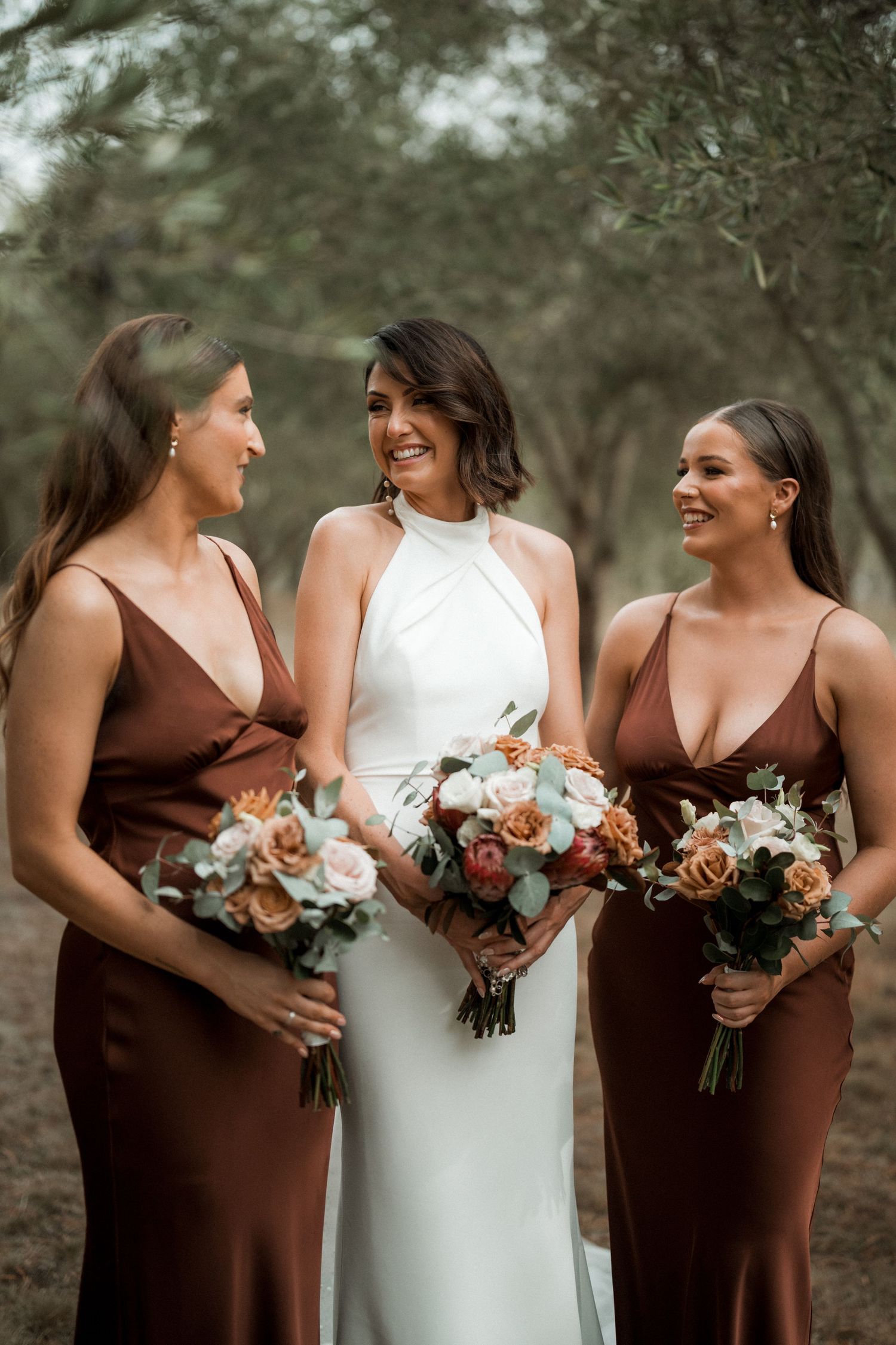 Three women in elegant brown and white dresses hold matching bouquets while sharing joyful smiles.