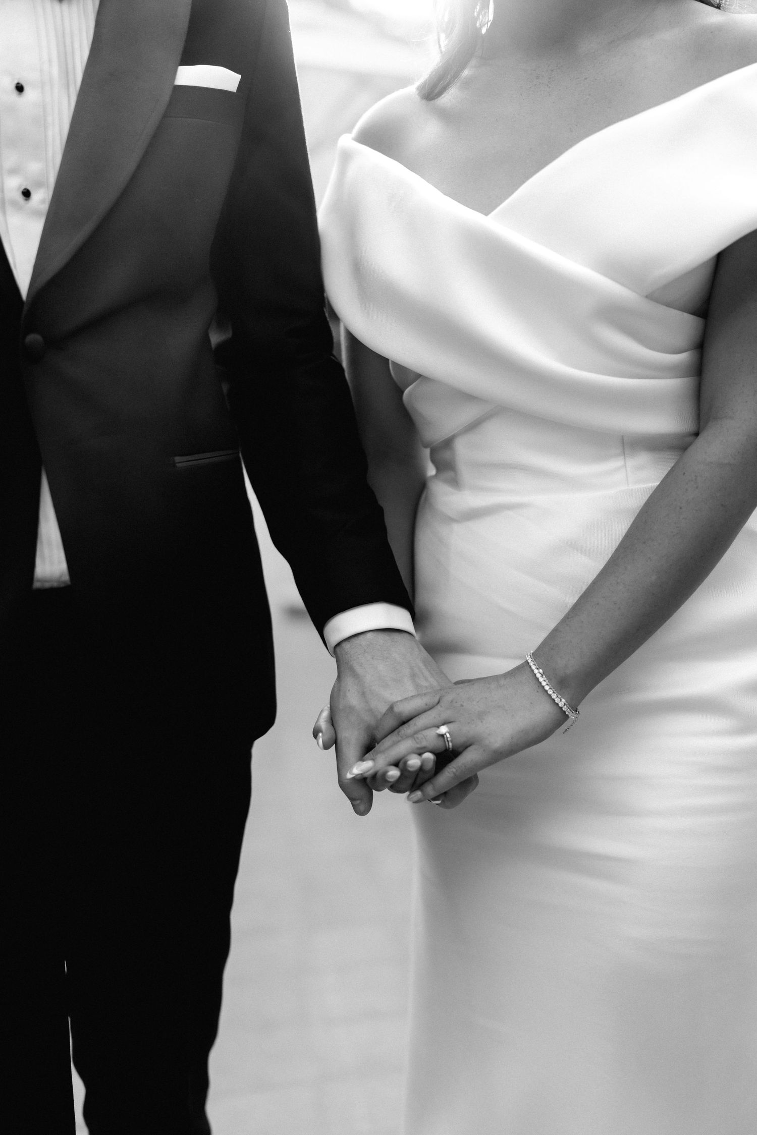 Black and white close-up of newlyweds holding hands, showing formal wedding attire and jewelry.