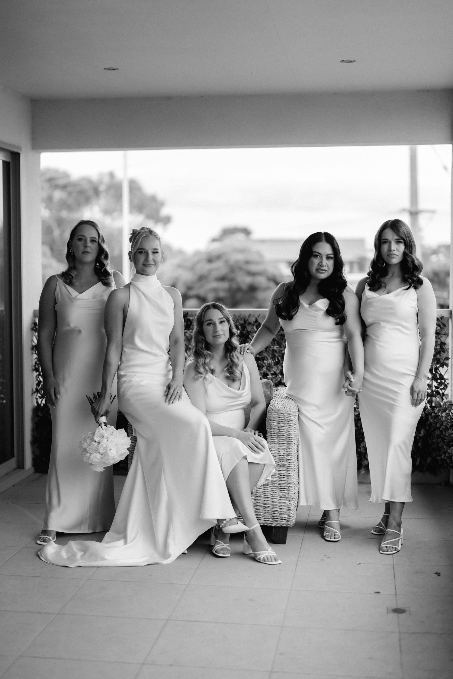 Five people in elegant white dresses pose together on a covered patio during a formal event.