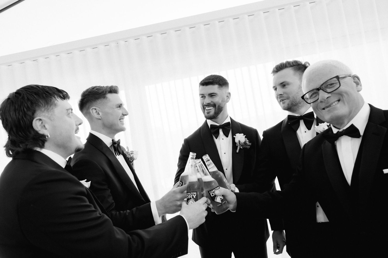 A group of groomsmen in black tuxedos share a candid moment of laughter during a wedding celebration.