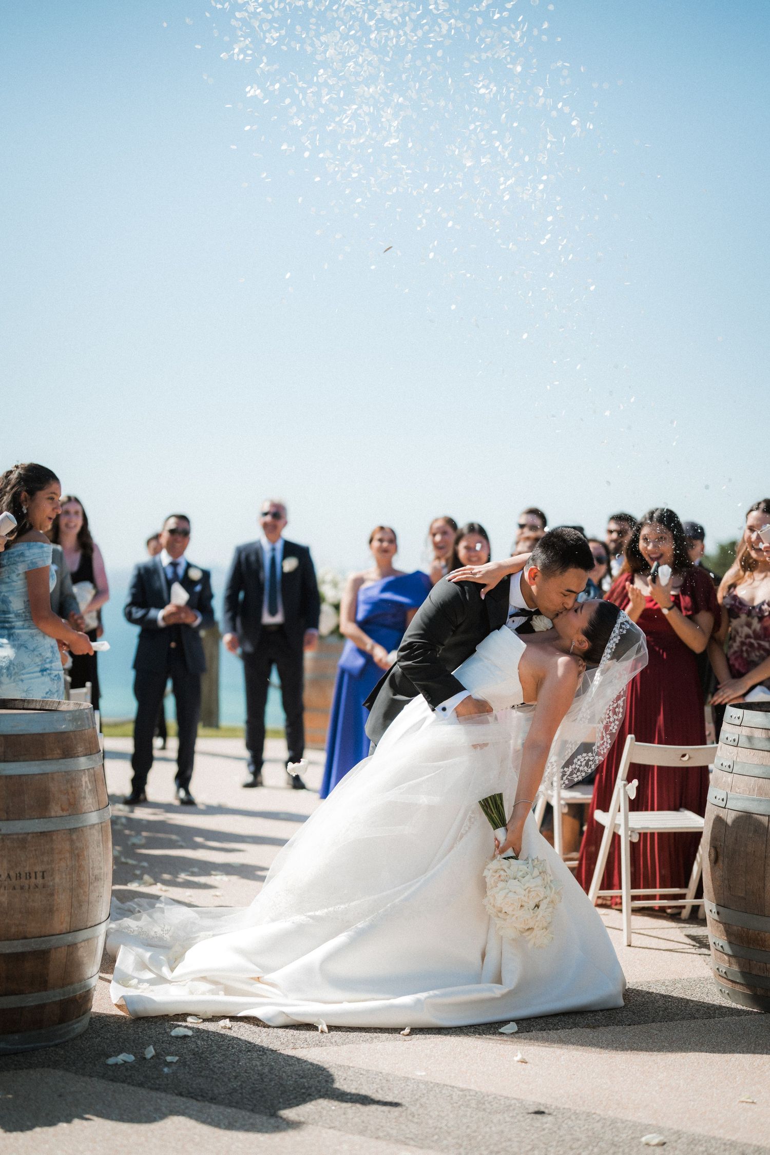Wedding guests celebrate as confetti falls during an outdoor ceremony near wine barrels.