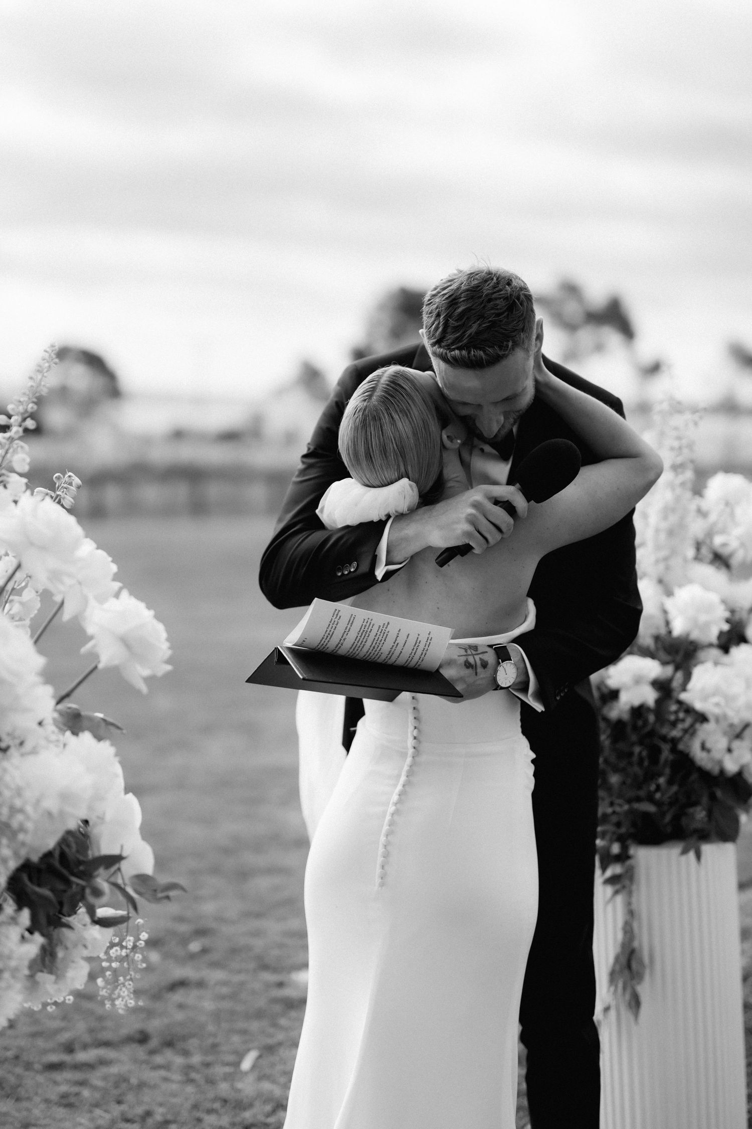 A tender embrace between a couple in formal wedding attire next to floral arrangements.