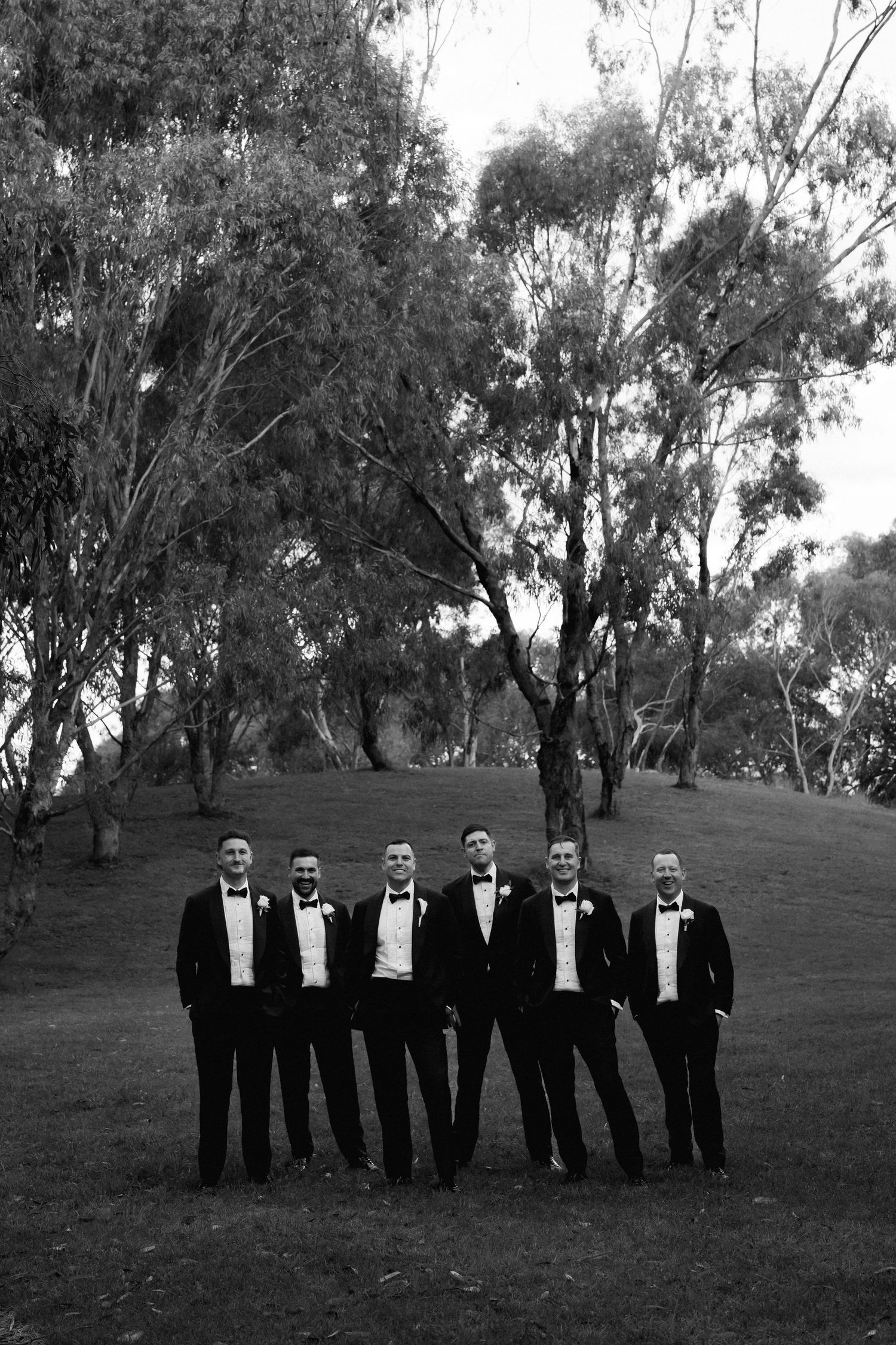 Groomsmen in black tuxedos pose together on a grassy hillside.