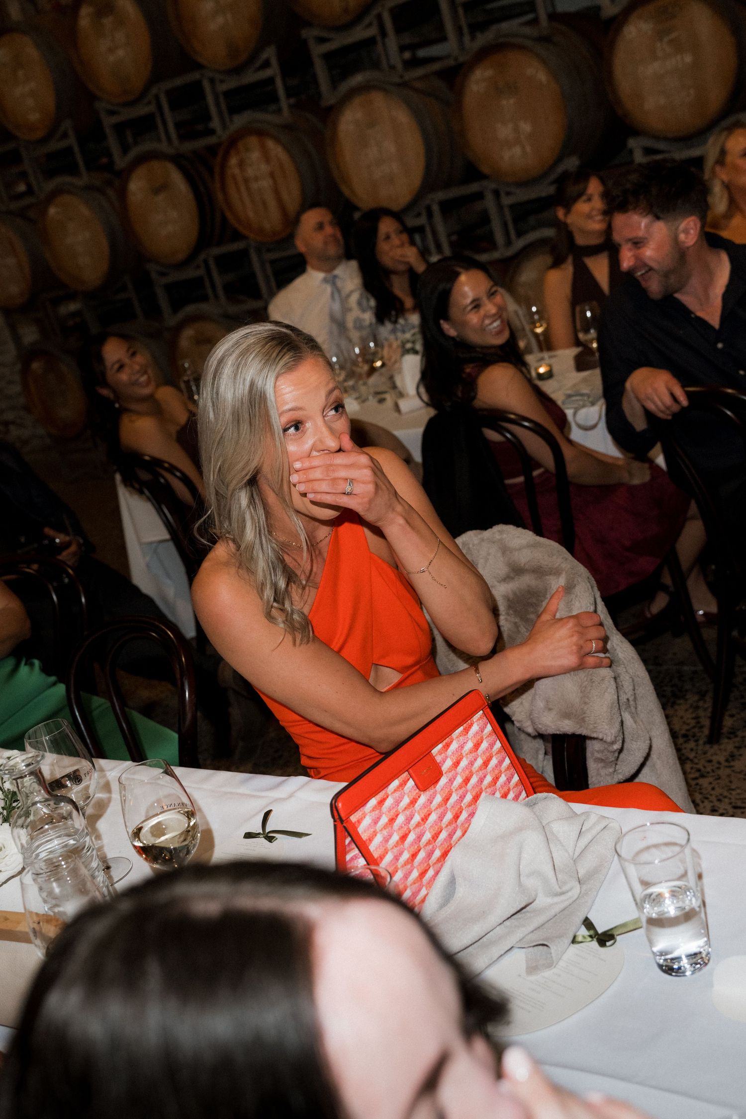 Wedding reception dinner scene with guests in orange attire seated at tables with wine barrels.