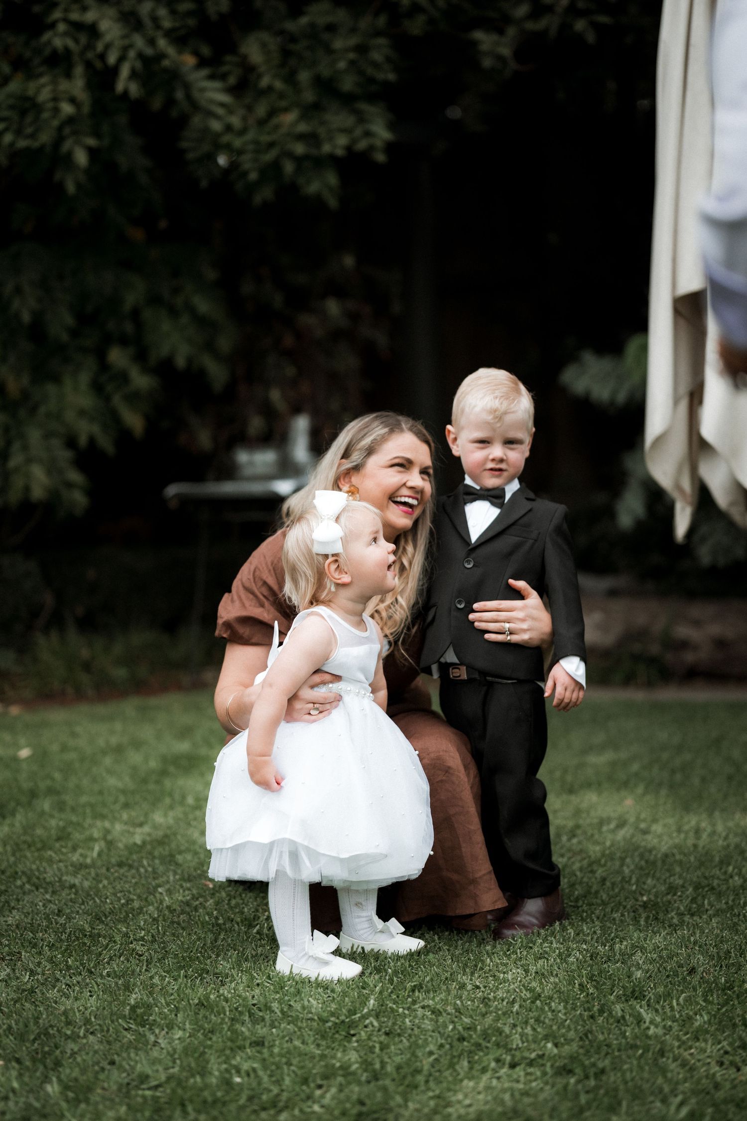 Children in formal wedding attire playing and sitting together on green grass during an outdoor celebration.
